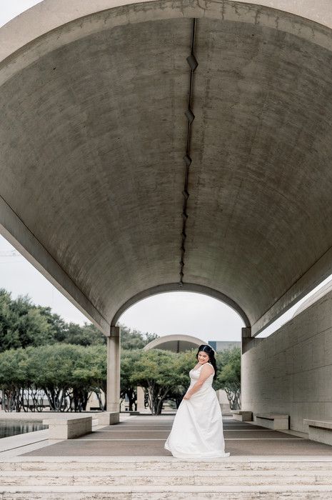Bride in white gown standing under curved concrete archway at modern architectural venue.