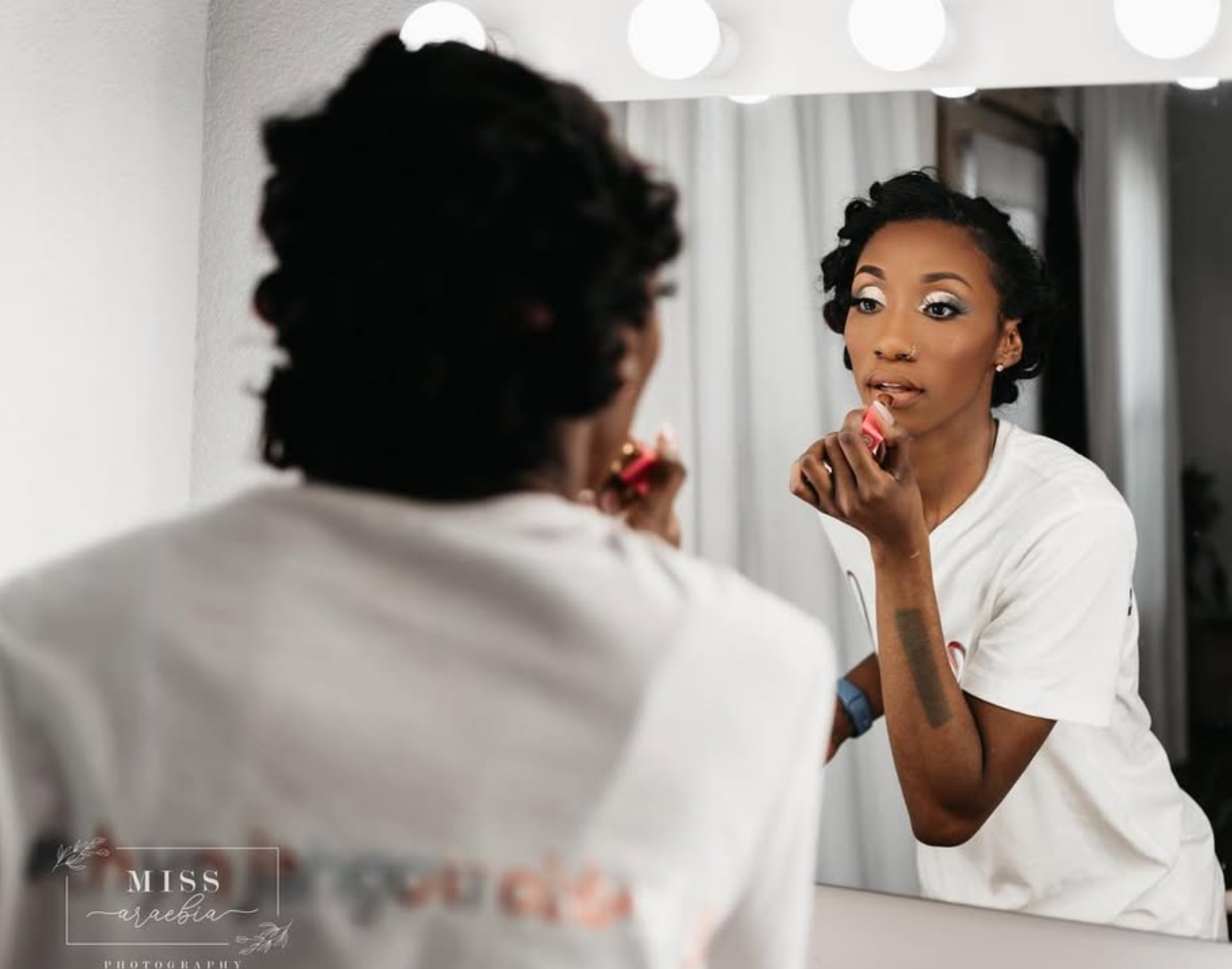 Young Black woman applying lipstick while looking in a lighted vanity mirror, wearing a white top.