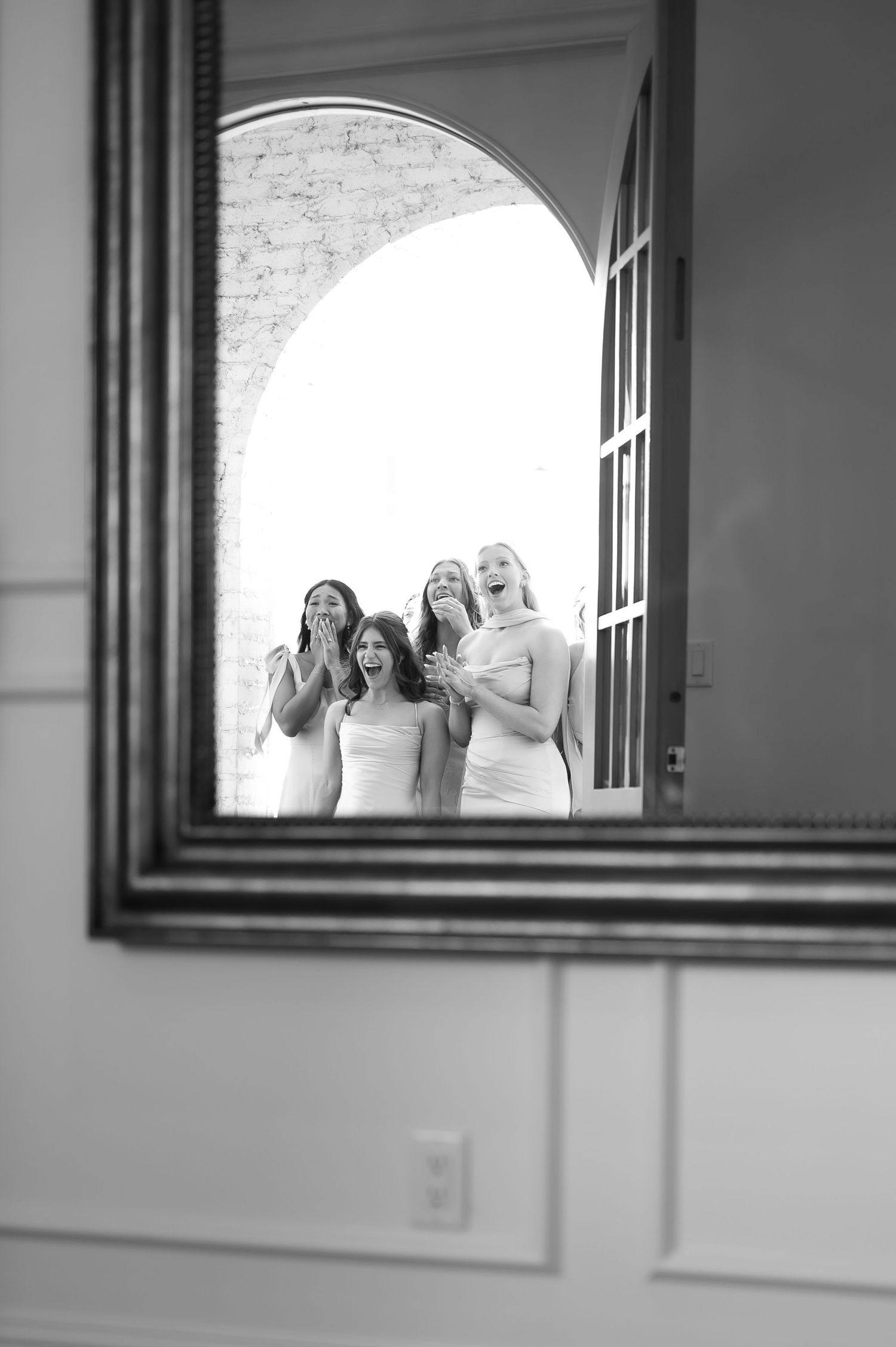 Four bridesmaids in white dresses laughing and posing together, captured in a vintage mirror reflection in black and white.