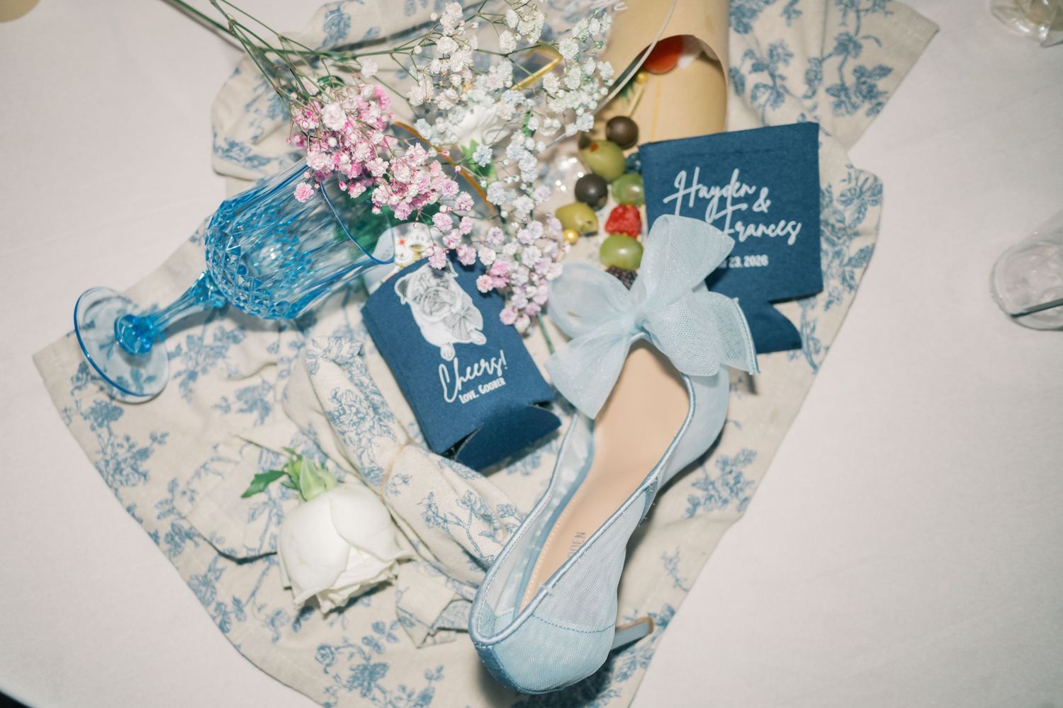 Flat lay of blue wedding accessories including shoes, koozies, floral bouquet, and toile napkin on a table.