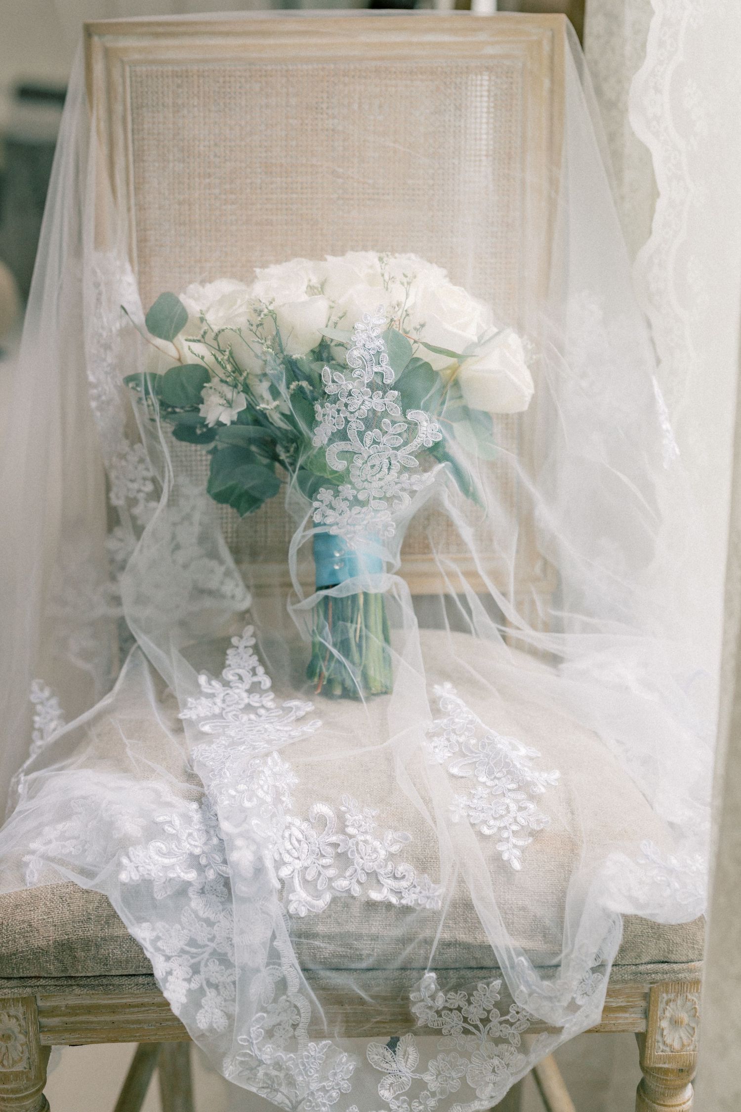 White rose wedding bouquet with eucalyptus draped in lace veil on vintage chair.
