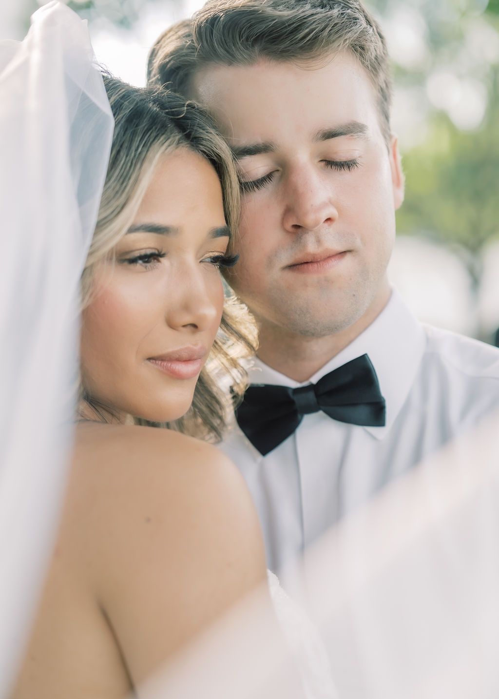 A bride and groom share a tender moment under a wedding veil, dressed in elegant white and black bow tie attire.