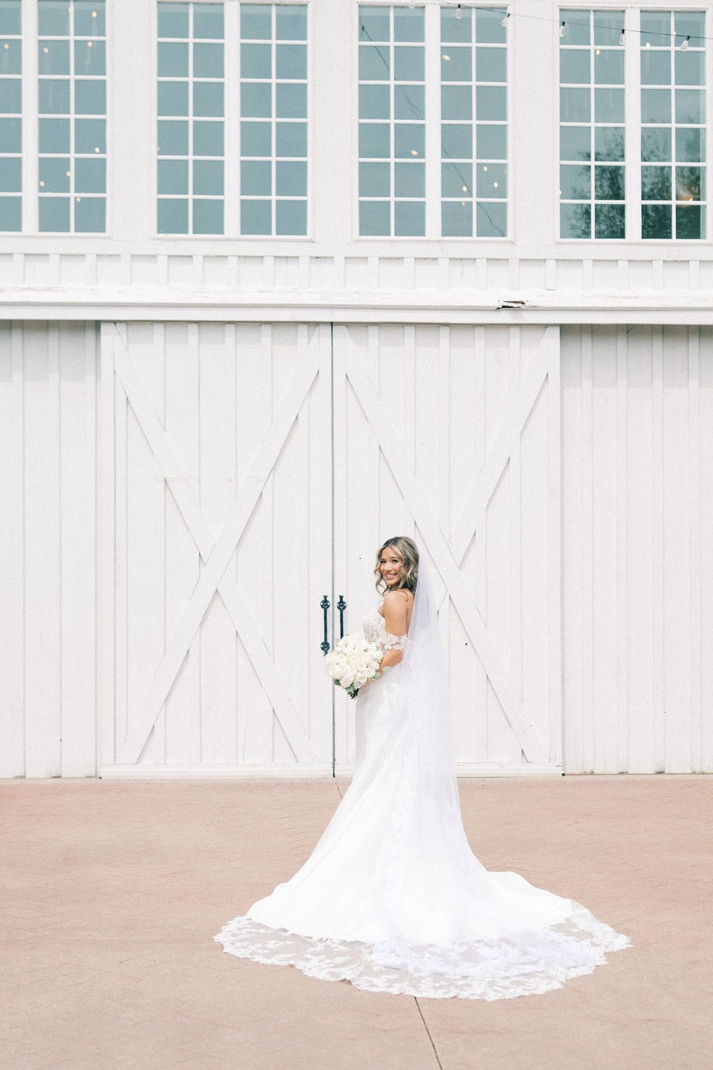 Bride in a white lace-trimmed gown with long veil holds bouquet, posing in front of white barn doors.
