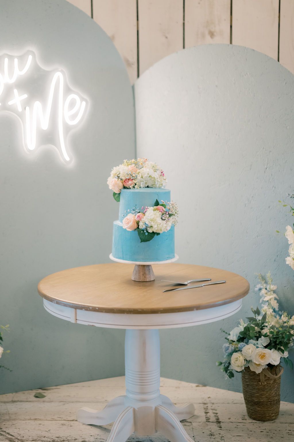Two-tier blue wedding cake adorned with pink and white flowers on a white pedestal table with neon sign backdrop.