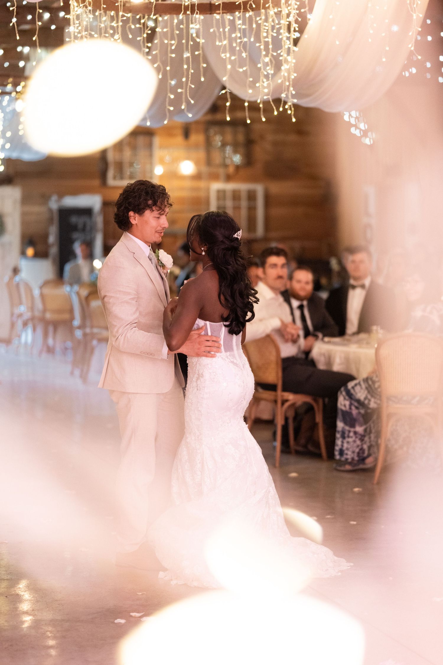 Newlyweds share romantic first dance under fairy lights and draped fabric at rustic barn reception.