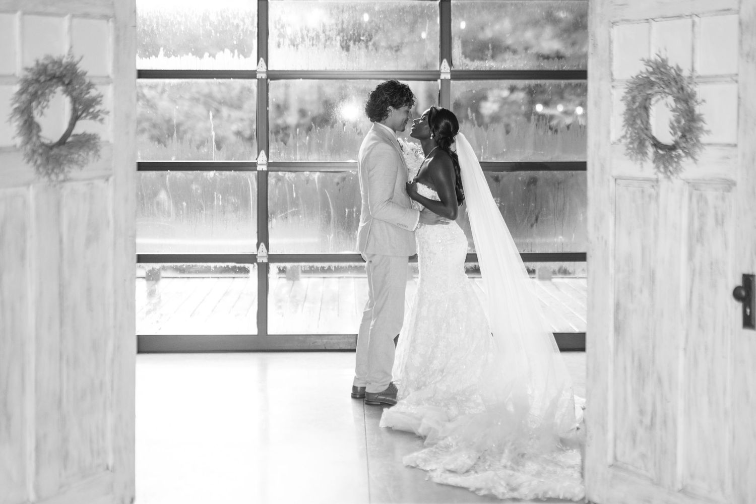 Black and white photo of bride and groom sharing first dance framed by open barn doors with wreaths.