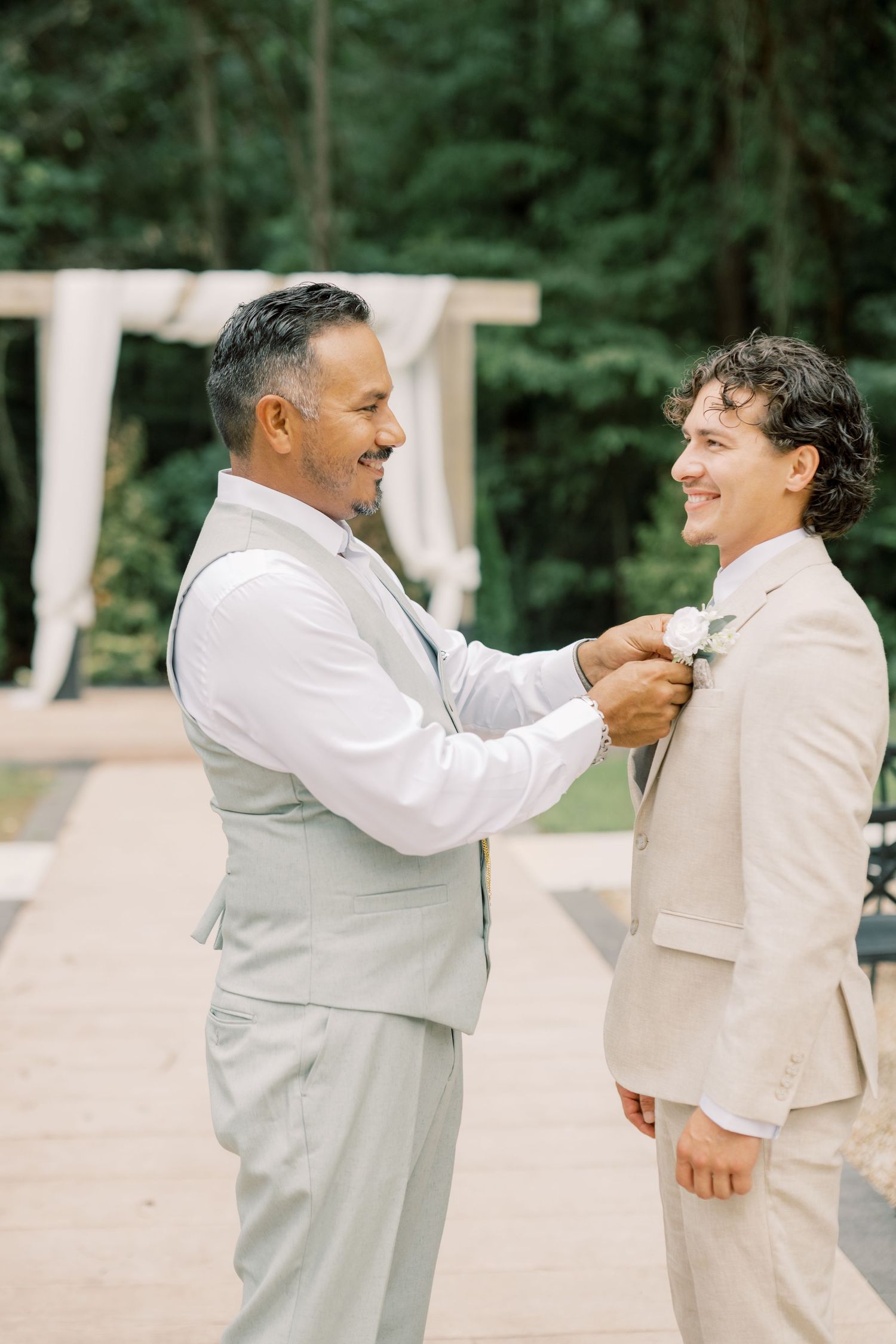 Two men in beige suits smile as one pins a white boutonniere on the other outdoors at a wedding ceremony.