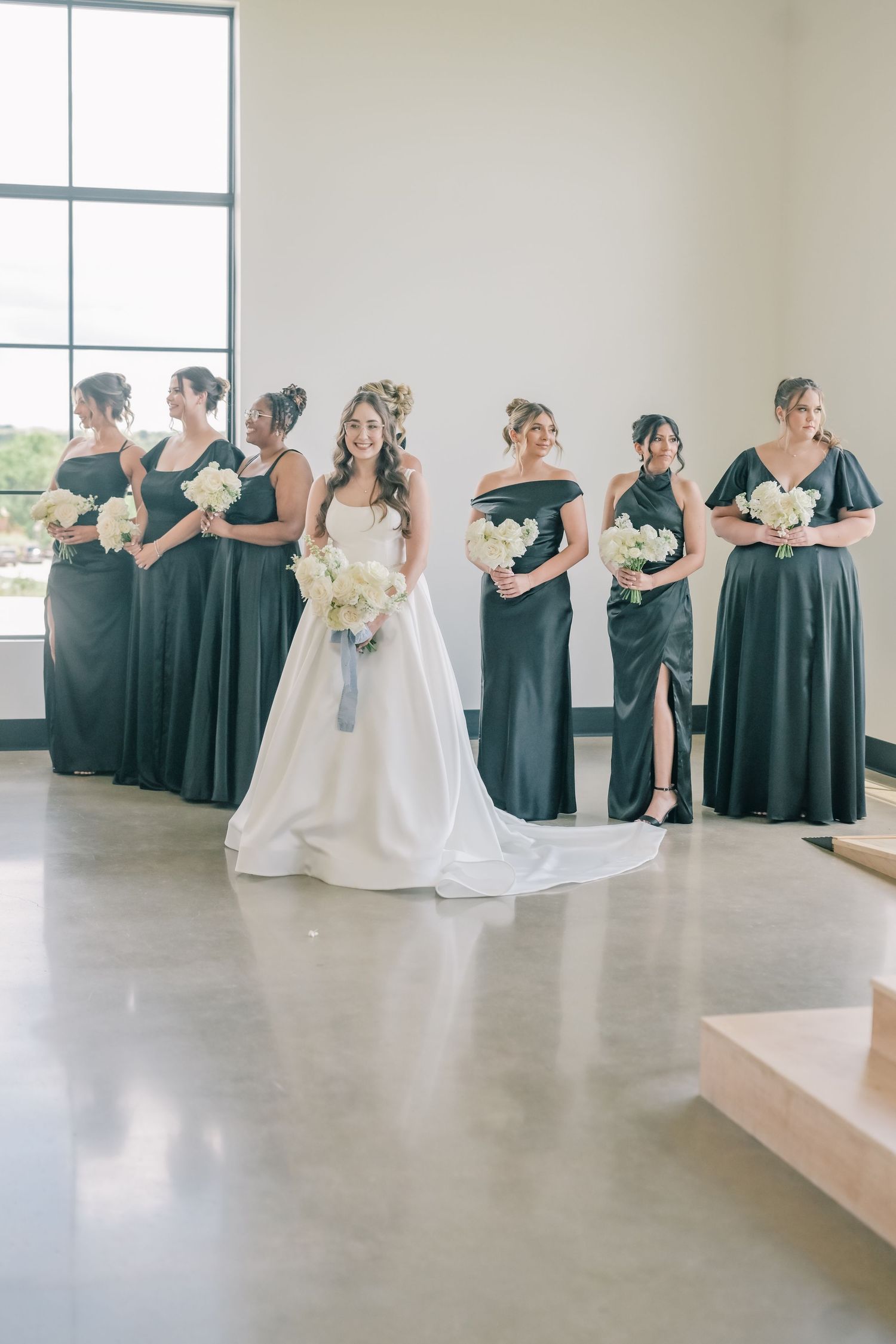 Bride in white ball gown stands with bridesmaids in dark green dresses holding white bouquets in elegant venue.