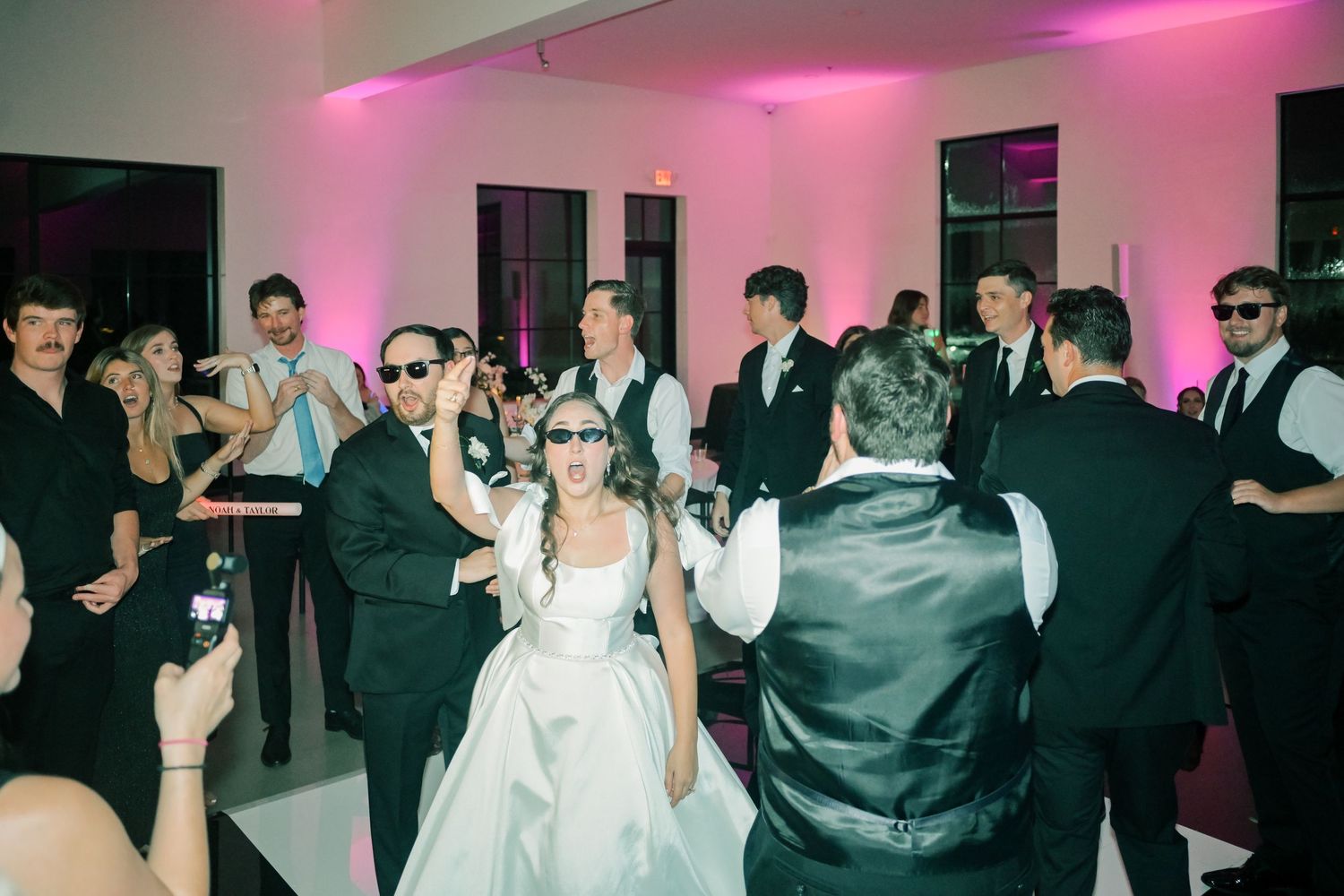 Bride and groom dancing with sunglasses-wearing wedding guests at lively reception with pink uplighting.