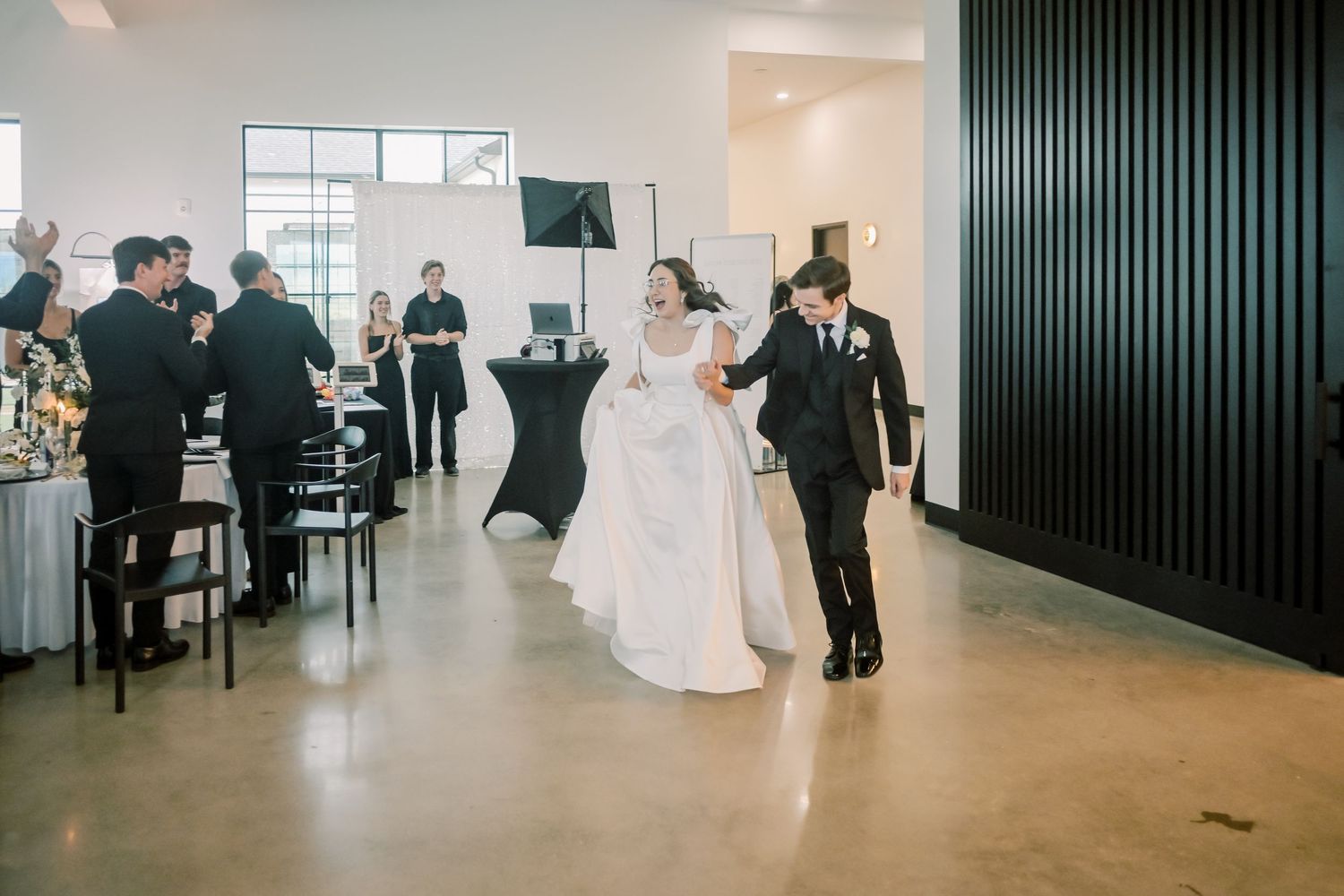 Bride and groom making grand entrance into modern wedding reception venue with guests applauding.