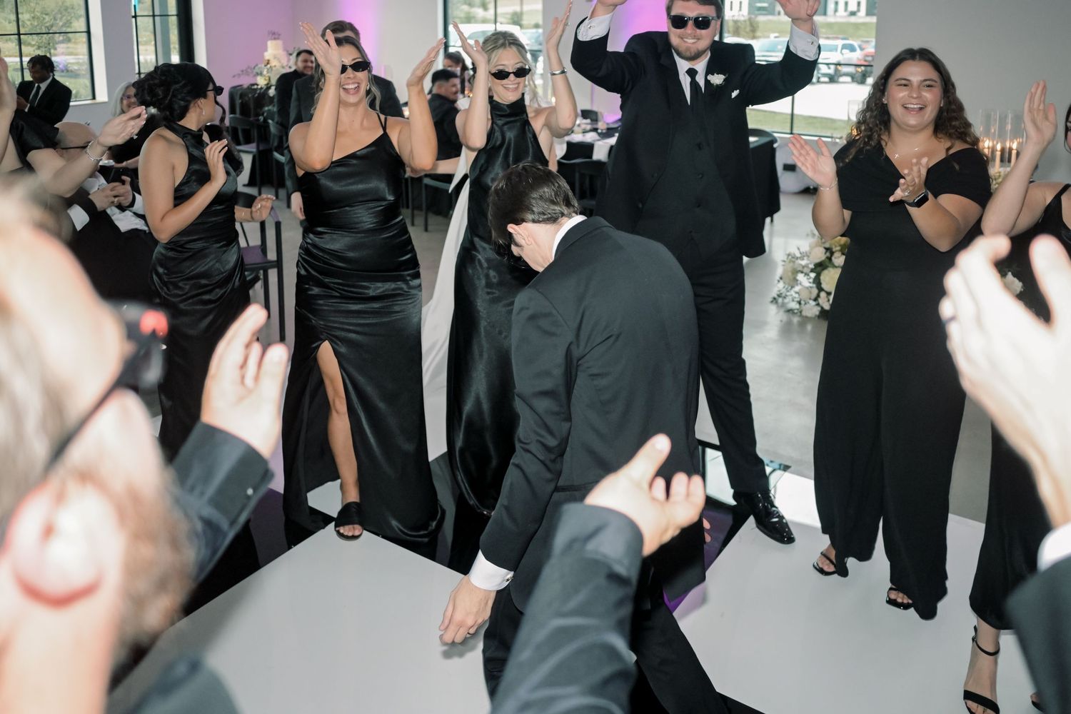 Wedding guests in black attire dancing energetically on a lit dance floor at a lively reception party.
