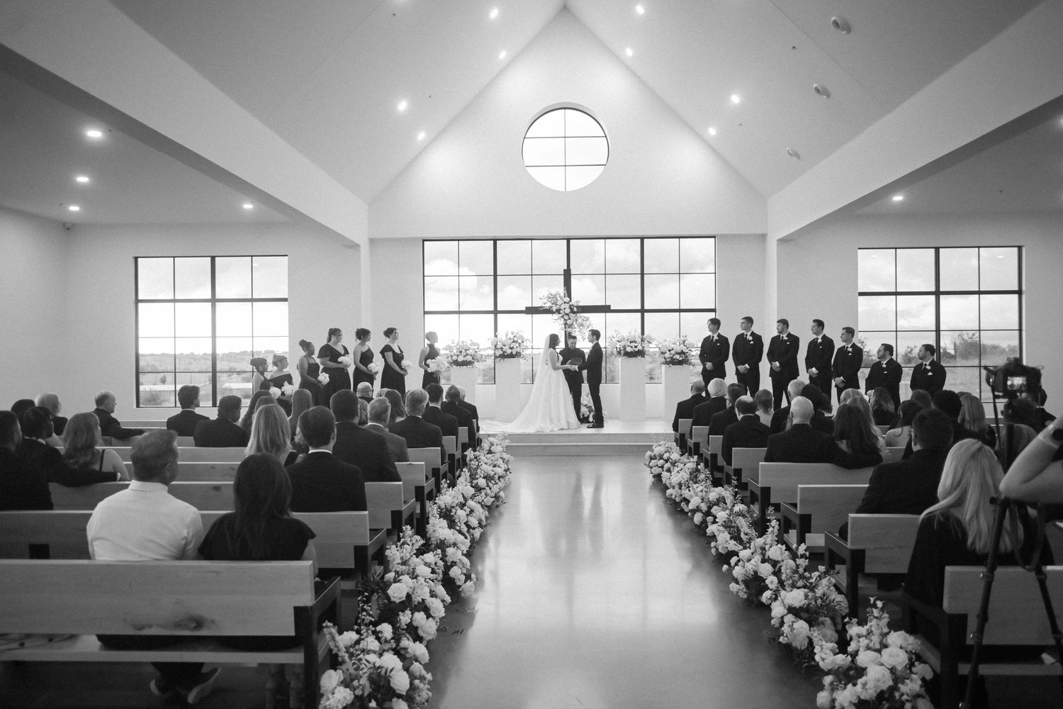 Black and white wide shot of wedding ceremony in bright chapel with floral aisle decorations.