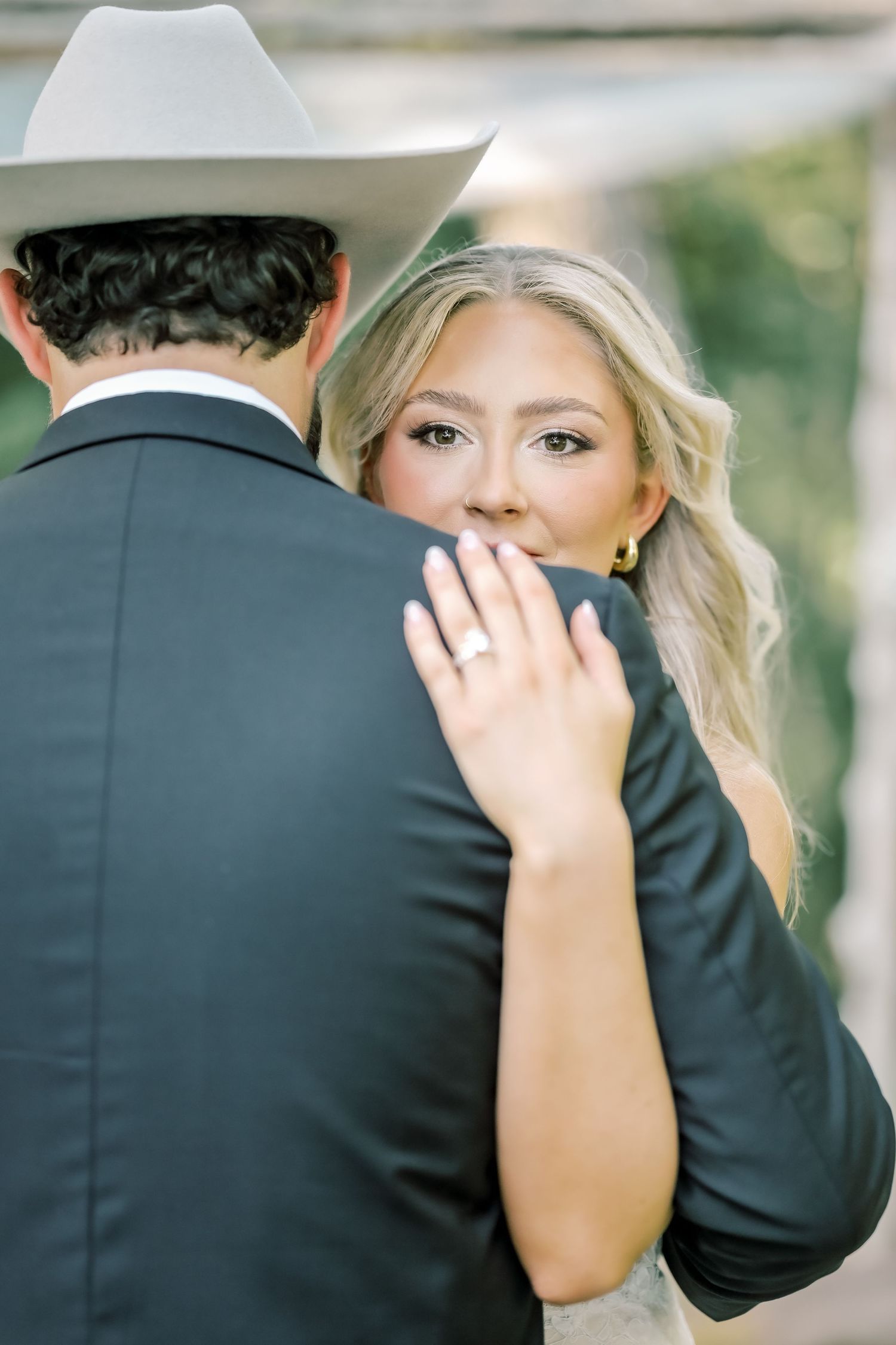 Bride in lace gown embraces groom in cowboy hat, showing diamond engagement ring.