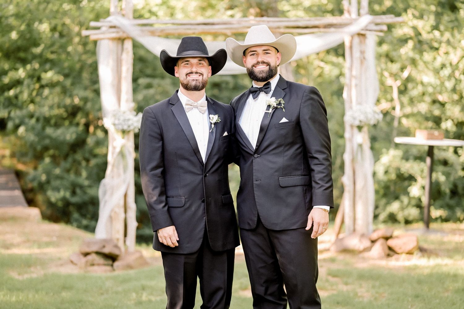 Two grooms in black suits and cowboy hats pose together outdoors at a rustic wedding ceremony.