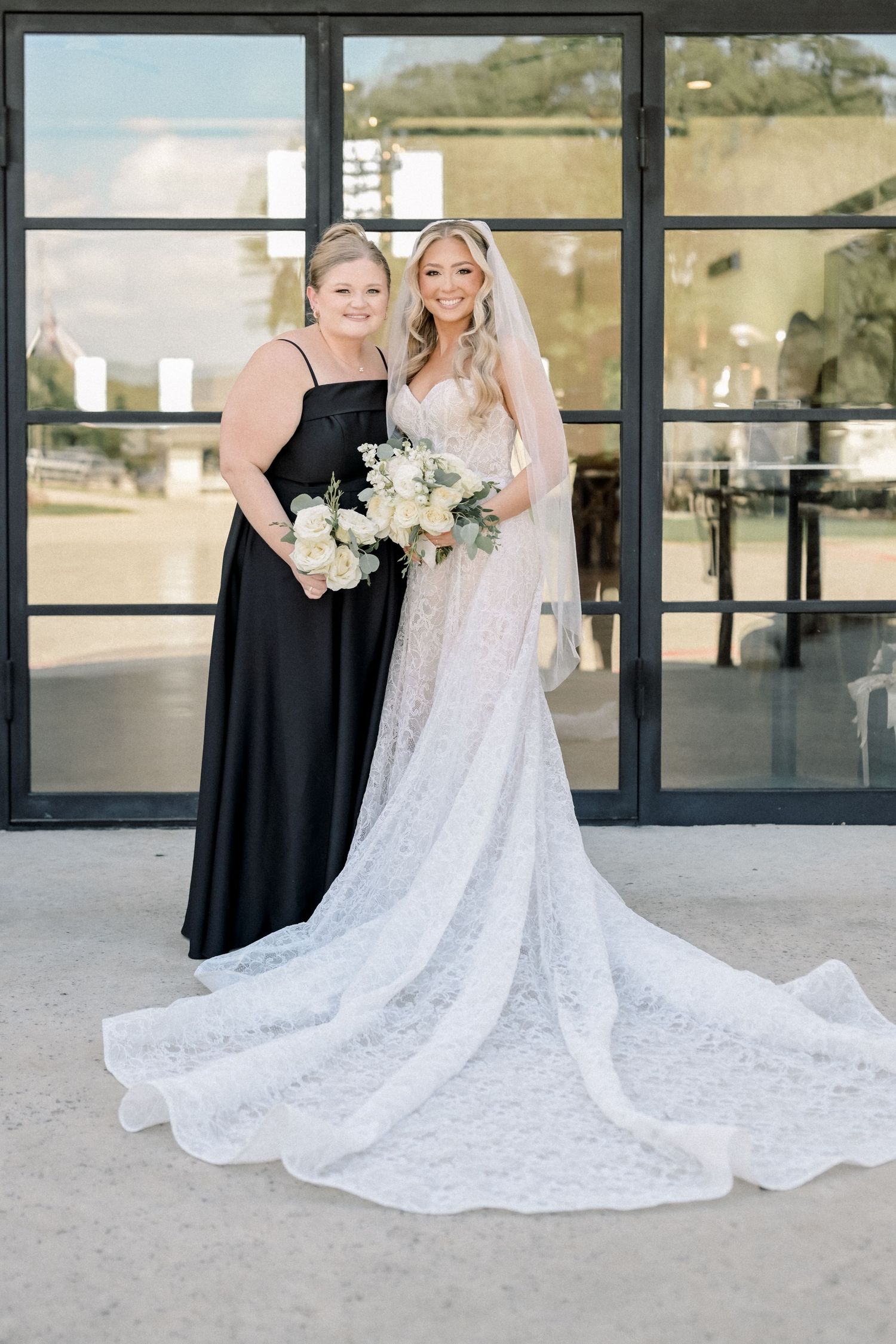 Bride in lace gown with long train poses with guest in black dress before large glass doors.