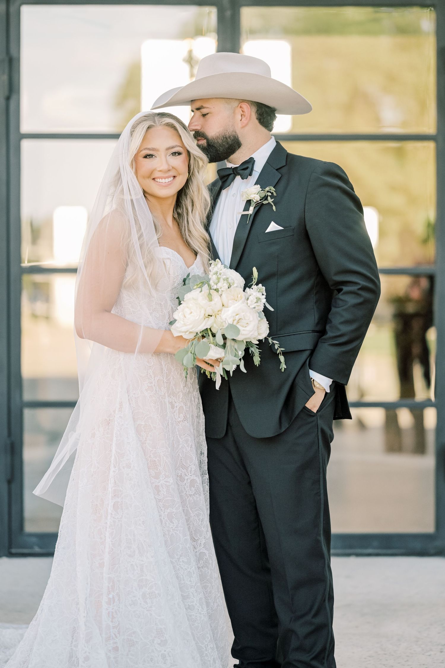 Bride and groom posing together, groom wearing cowboy hat, bride holding white floral bouquet.
