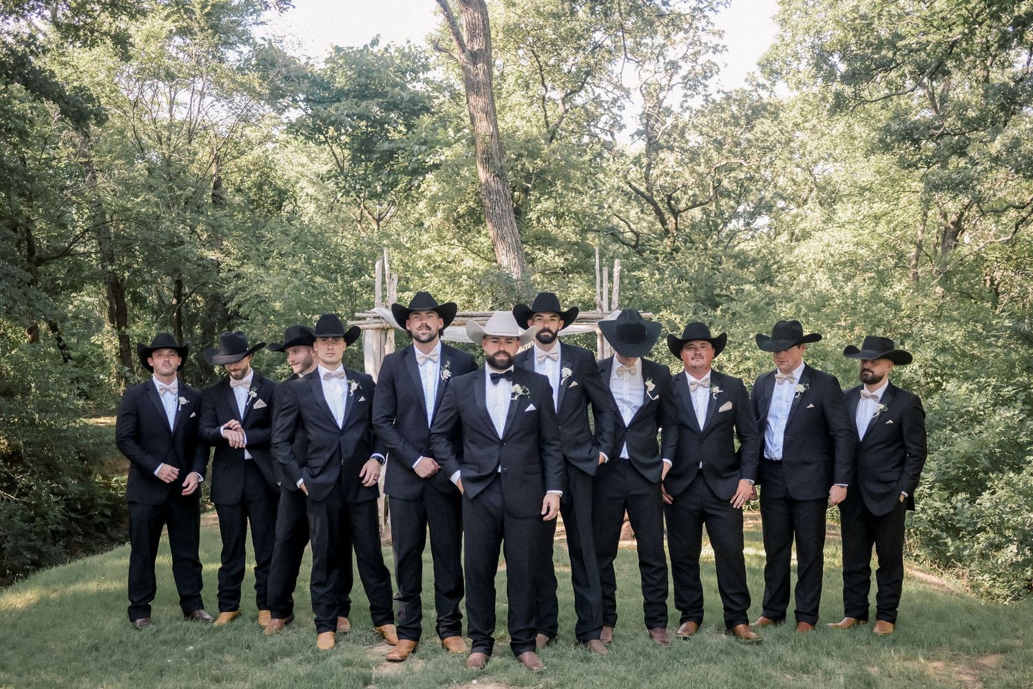 Groom and groomsmen in black suits and cowboy hats pose together in wooded outdoor wedding setting.