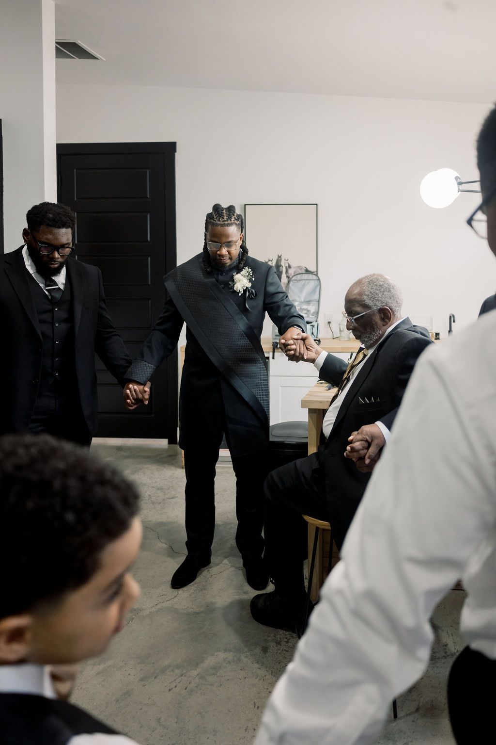 Groomsmen in black suits holding hands in prayer circle before wedding ceremony indoors.