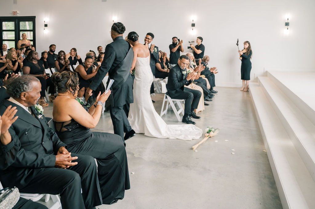 Wedding couple walks down the aisle as guests in black attire cheer and applaud in a bright white venue.