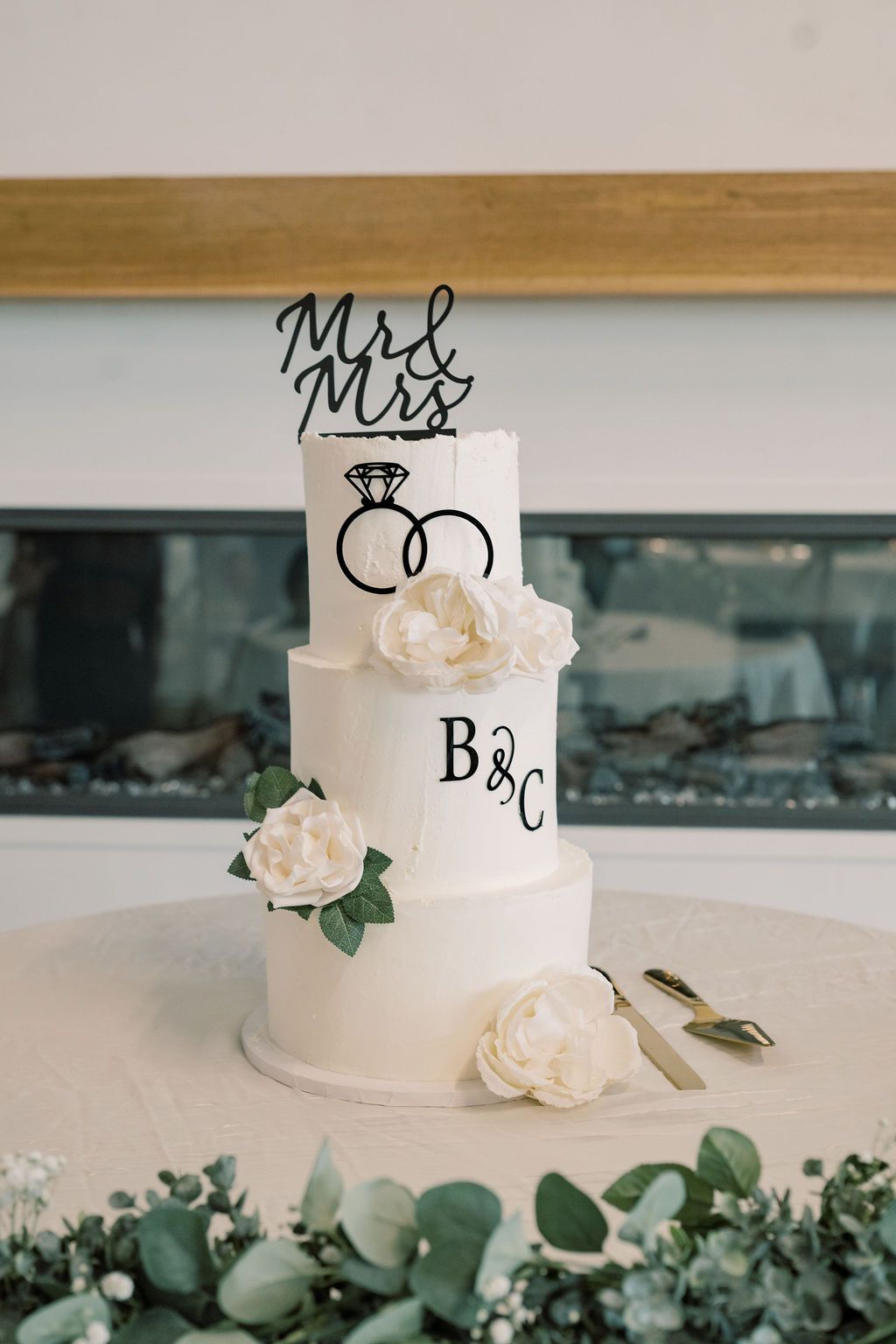 Elegant three-tier white wedding cake with Mr & Mrs topper, intertwined rings, B&C monogram, and white peony flowers.