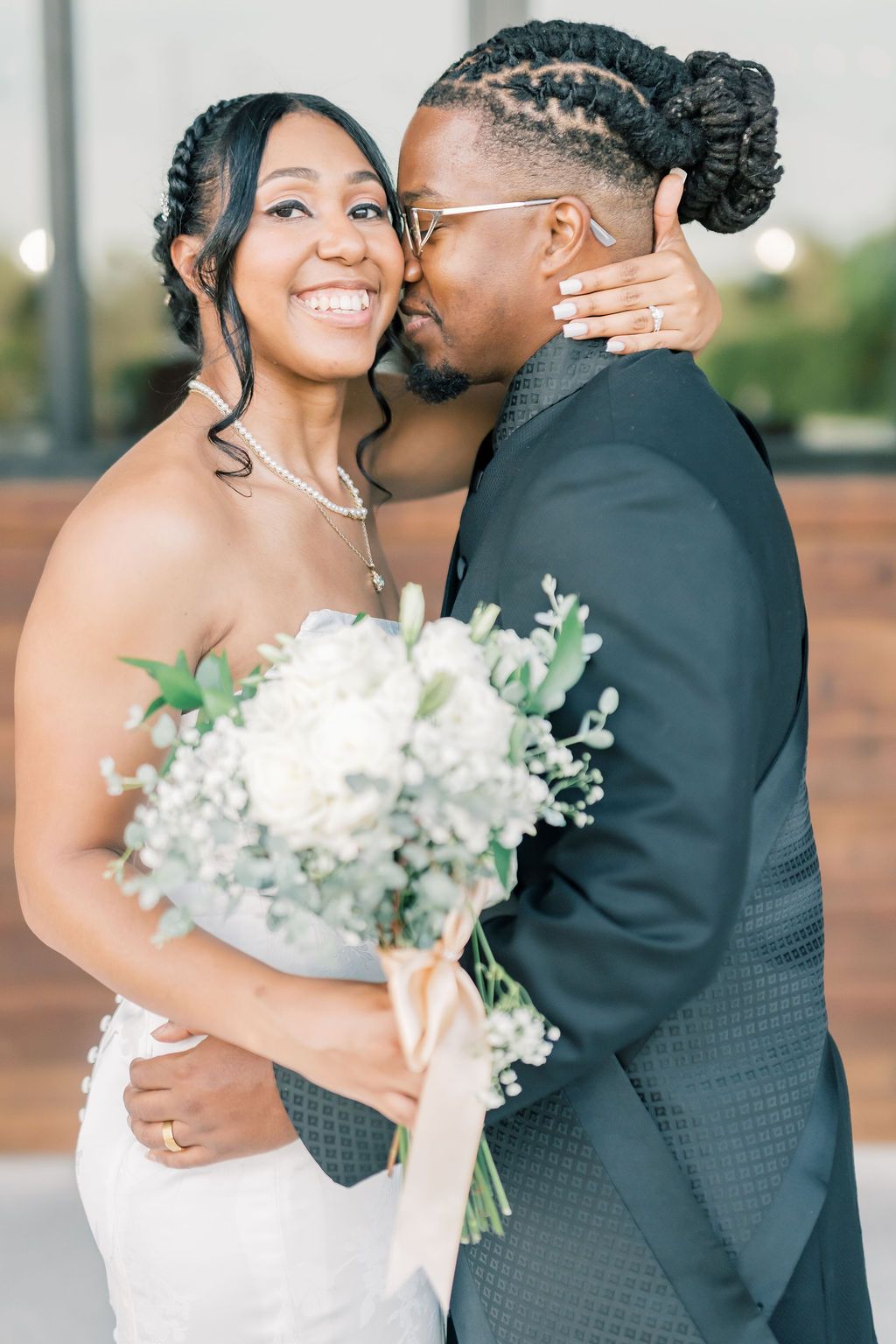A joyful bride holds white flowers while her groom kisses her cheek during their wedding day portrait.