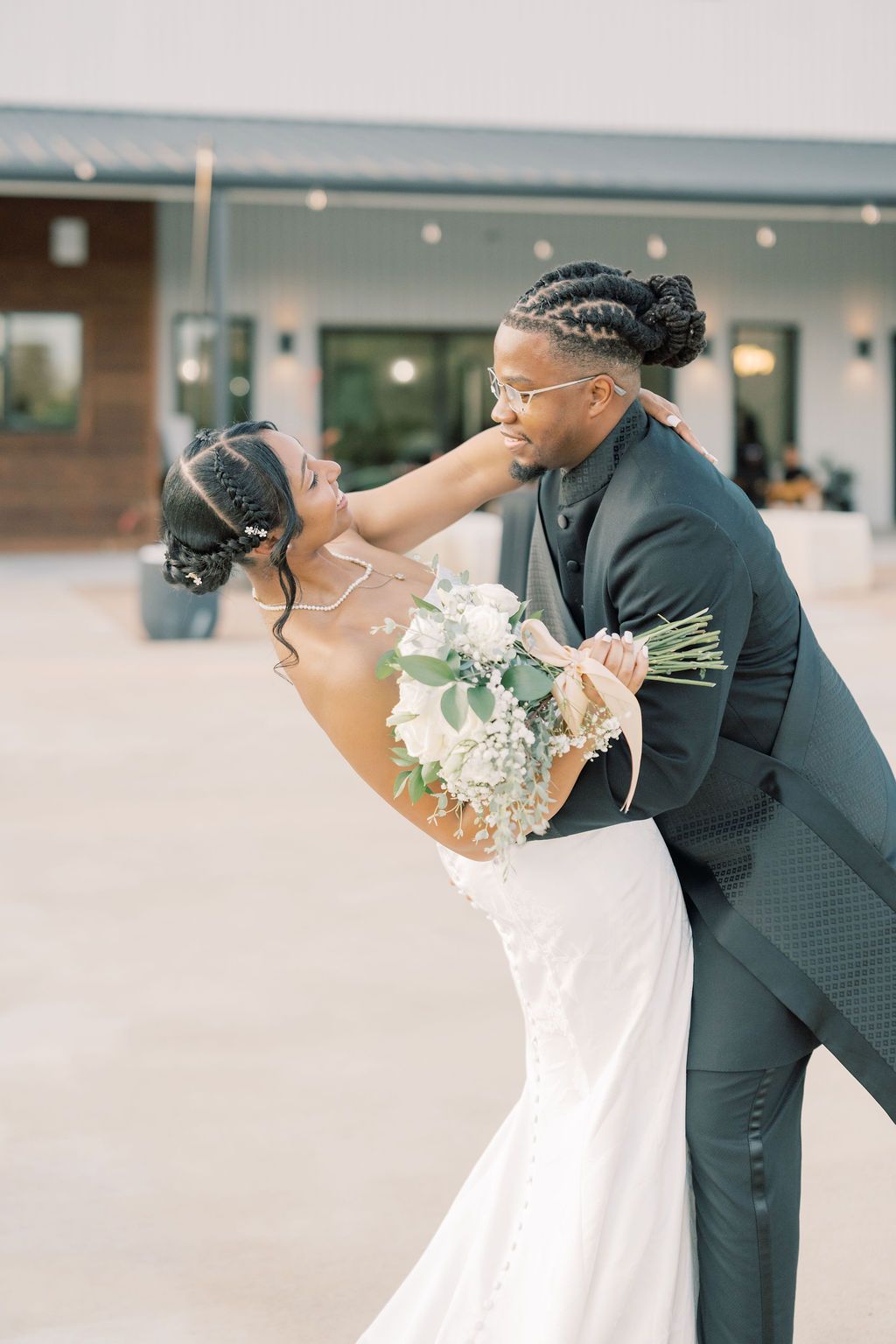Groom dips bride in romantic pose as she holds white floral bouquet at outdoor wedding.