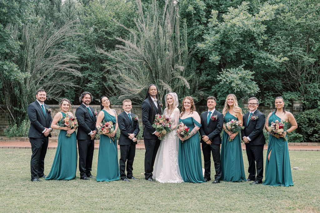 Wedding party in teal gowns and dark suits poses outdoors with lush green trees, bride in white holding floral bouquet.