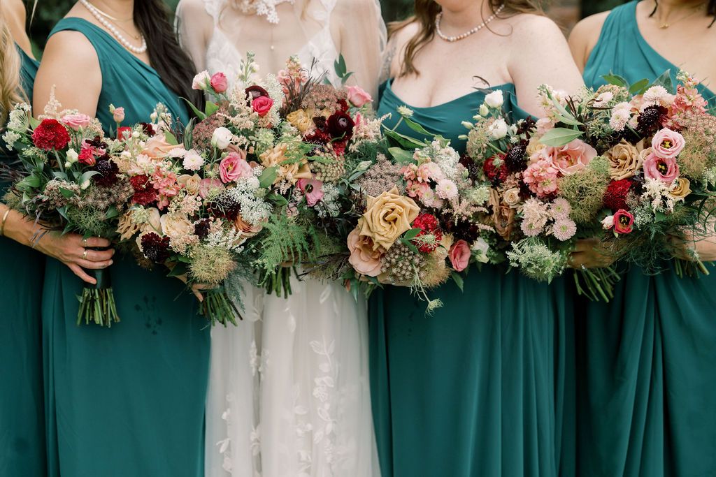 Bridesmaids in teal gowns and bride in white holding lush floral bouquets with red, pink, and cream blooms.