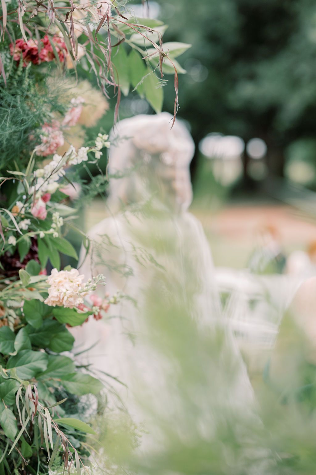 Lush floral arch with greenery and pink blooms frames a blurred outdoor wedding ceremony in a garden setting.
