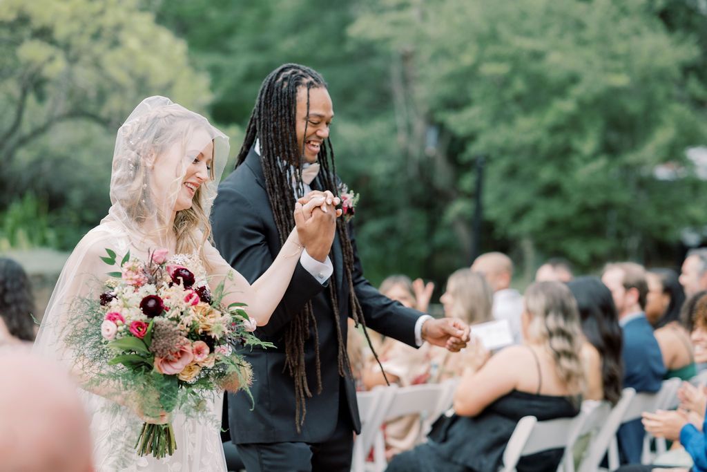 Joyful newlywed couple walking down the aisle, bride holding colorful bouquet, groom in black suit with dreadlocks.