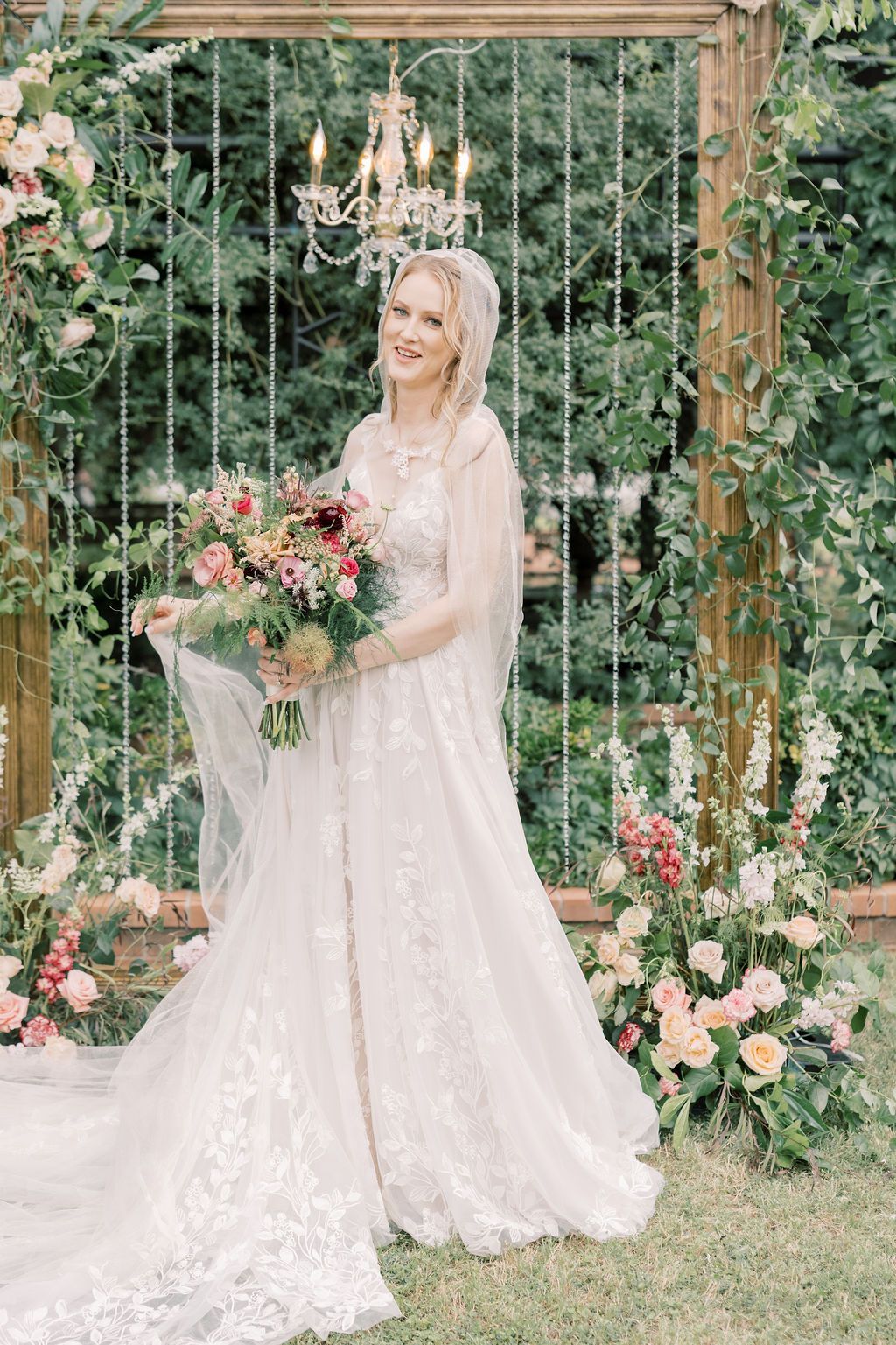 Smiling bride in lace gown and veil holds floral bouquet before a floral arch with chandelier outdoors.