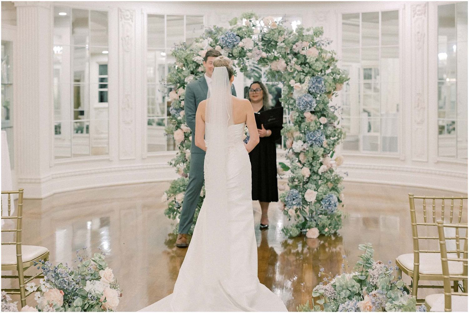 Wedding ceremony space decorated with lush blue and white floral arch in an elegant white ballroom.