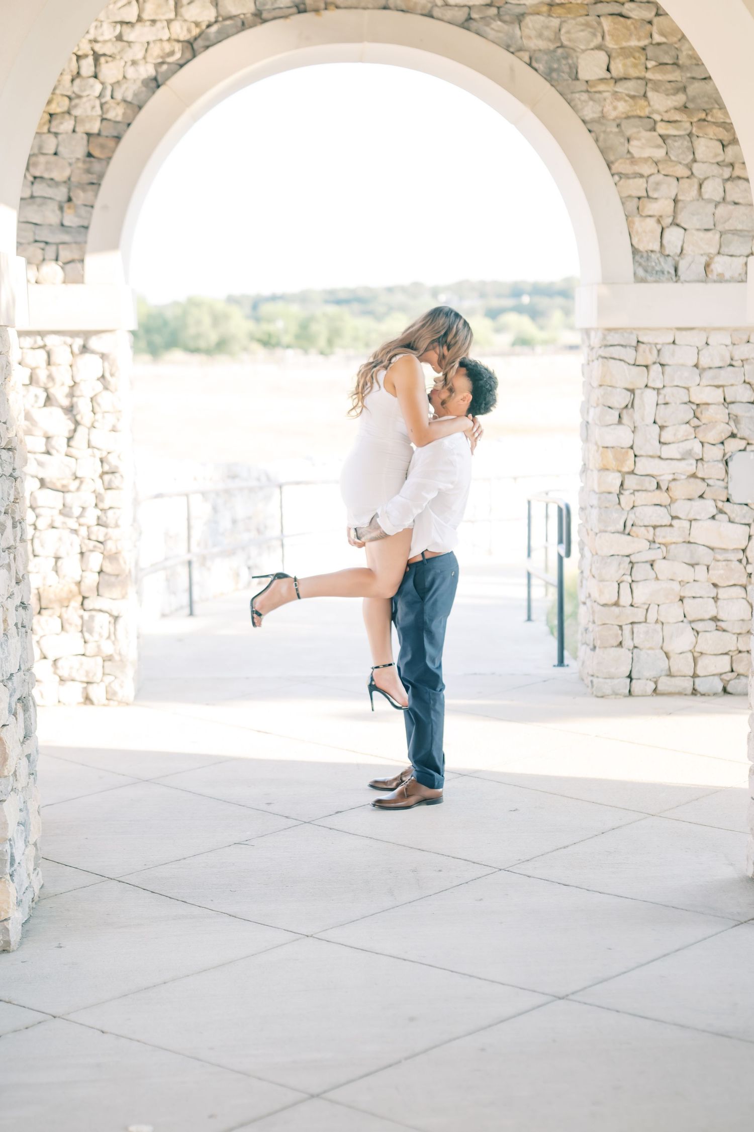 Man lifting woman for a kiss under elegant stone arch during engagement photos.