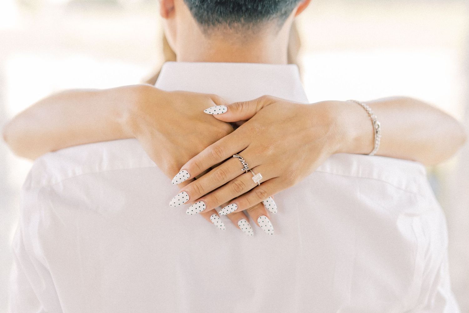 Woman with white floral nail art and diamond ring embracing man in white shirt from behind.