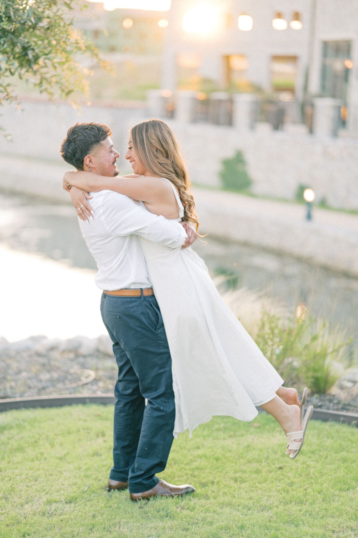 Man lifting woman in white dress during sunset engagement photos near a waterway.