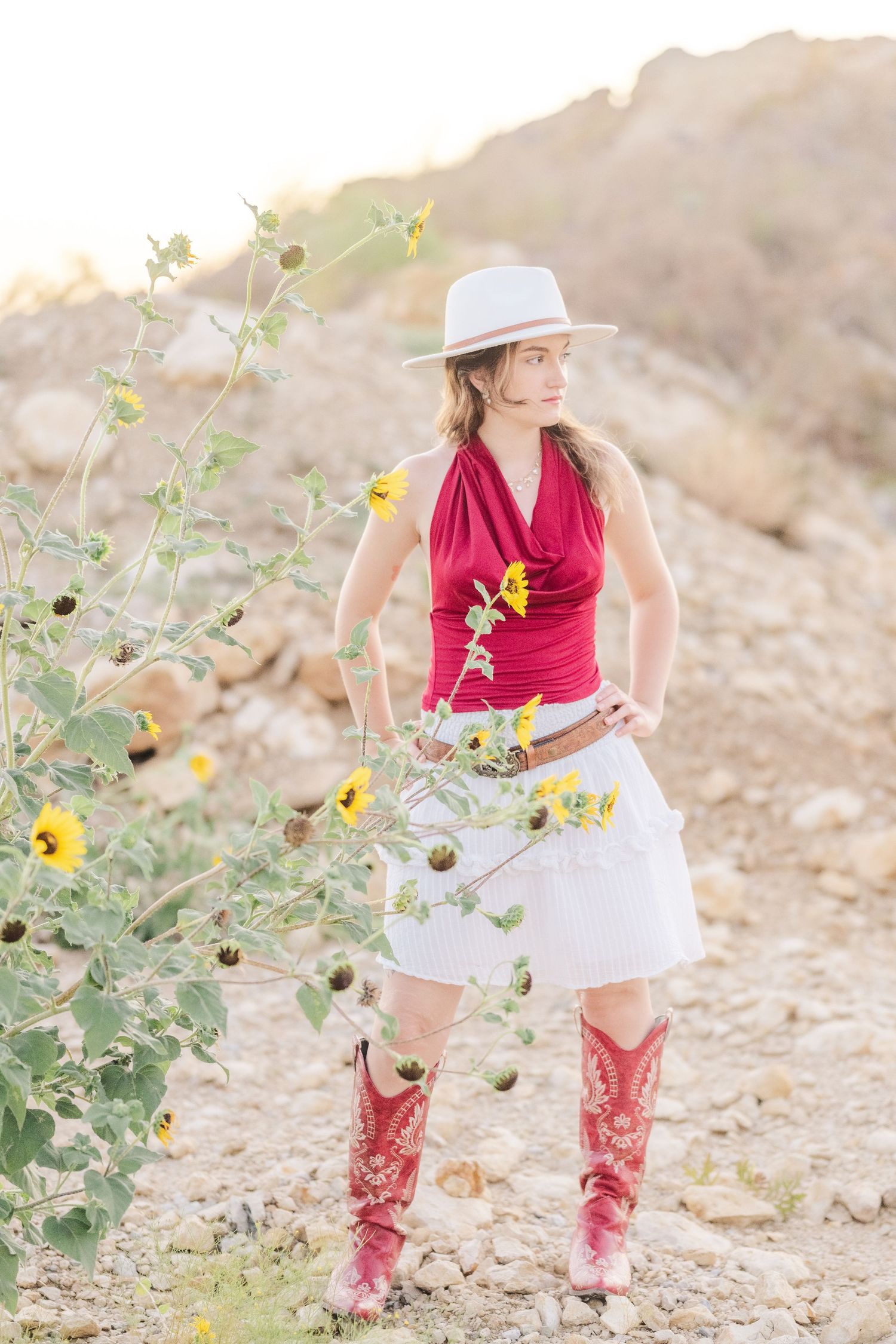 Woman in red halter top, white skirt, red cowboy boots and white hat stands among wild sunflowers in rocky desert.