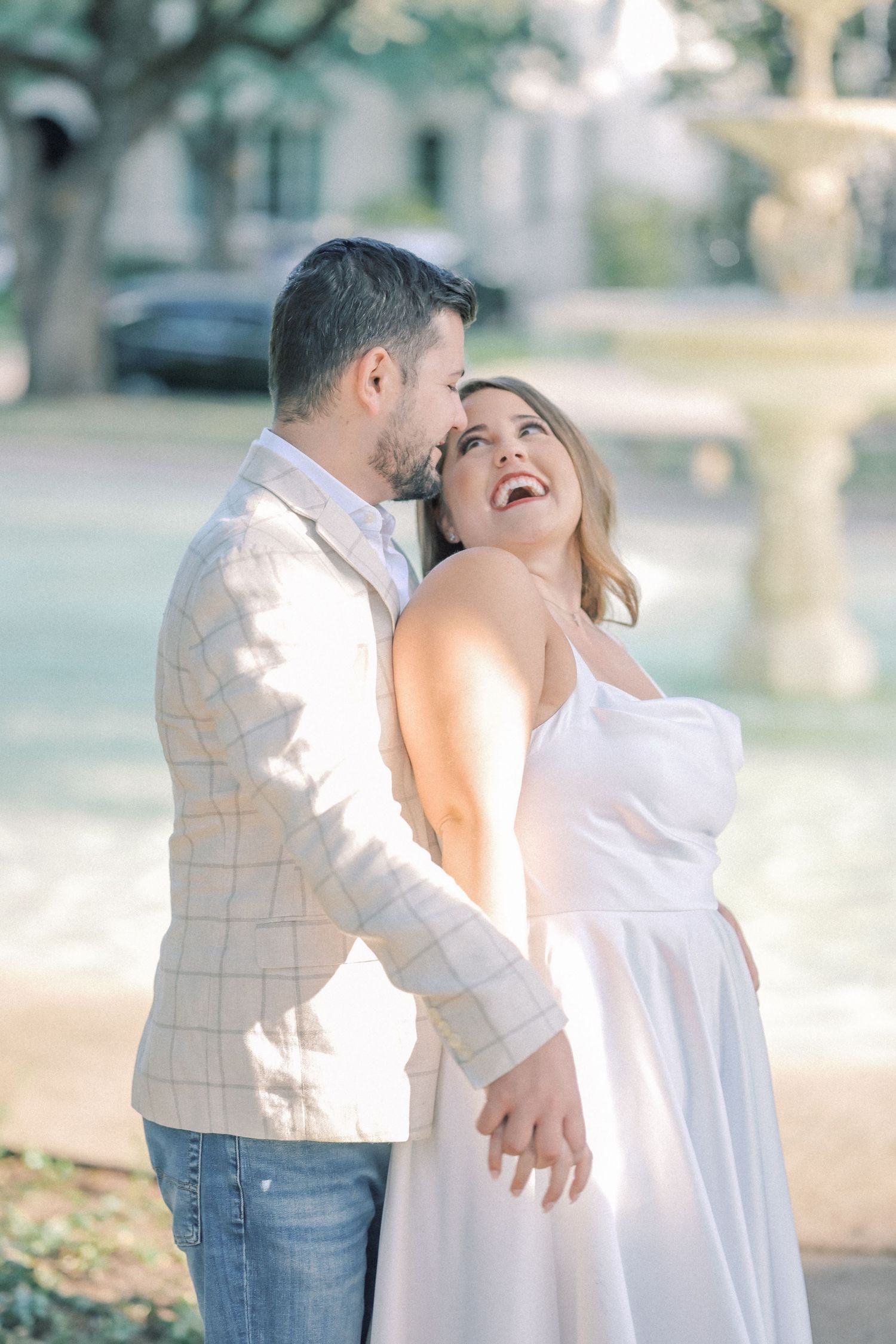 Smiling woman in a white dress laughs as a bearded man in a plaid blazer nuzzles her head near a stone fountain.