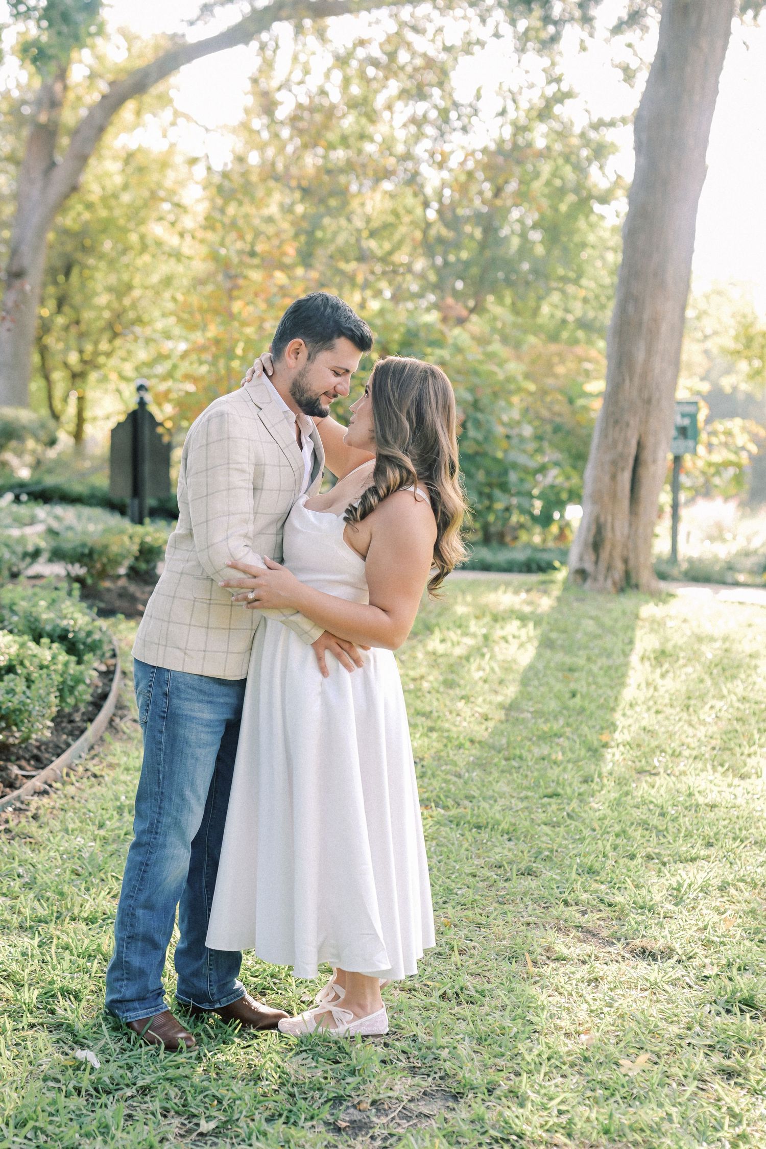 Couple sharing a tender embrace on a sunlit grassy lawn beneath large trees during an autumn engagement photo session.