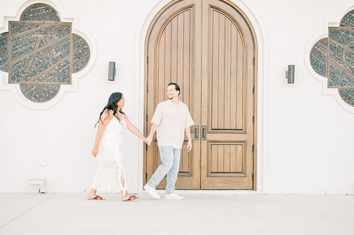A couple in white and blue outfits poses in front of an ornate wooden door for wedding photos.