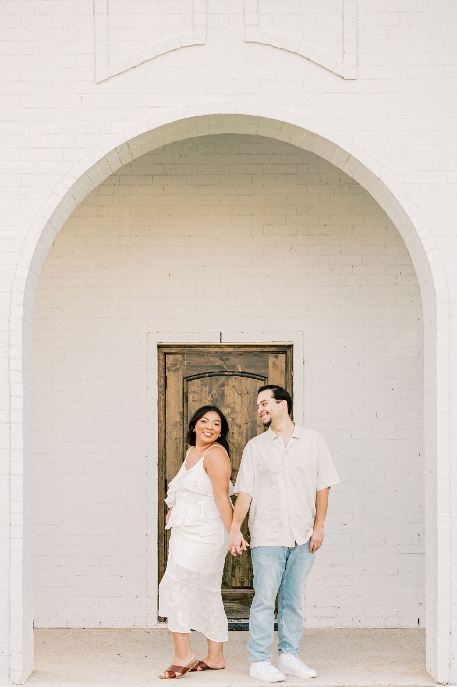 Two people stand together in a minimalist white arched hallway with vintage wooden doors.