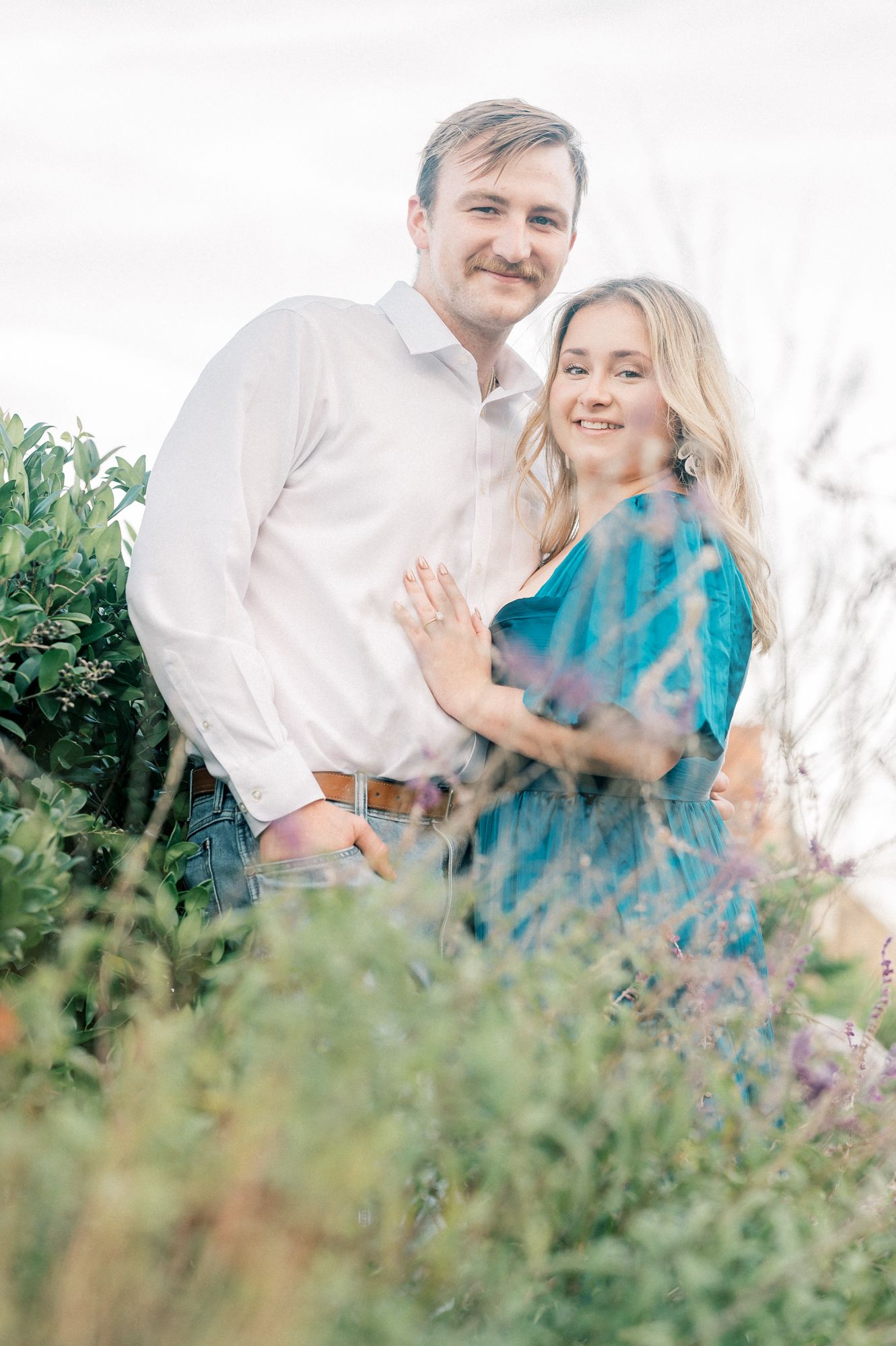 A romantic couple poses together in a field of wildflowers and tall grass on a sunny day.