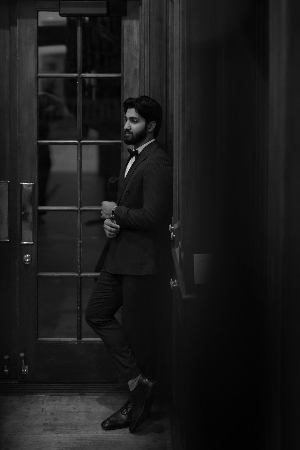 Moody black and white portrait of man in suit leaning against window frame.