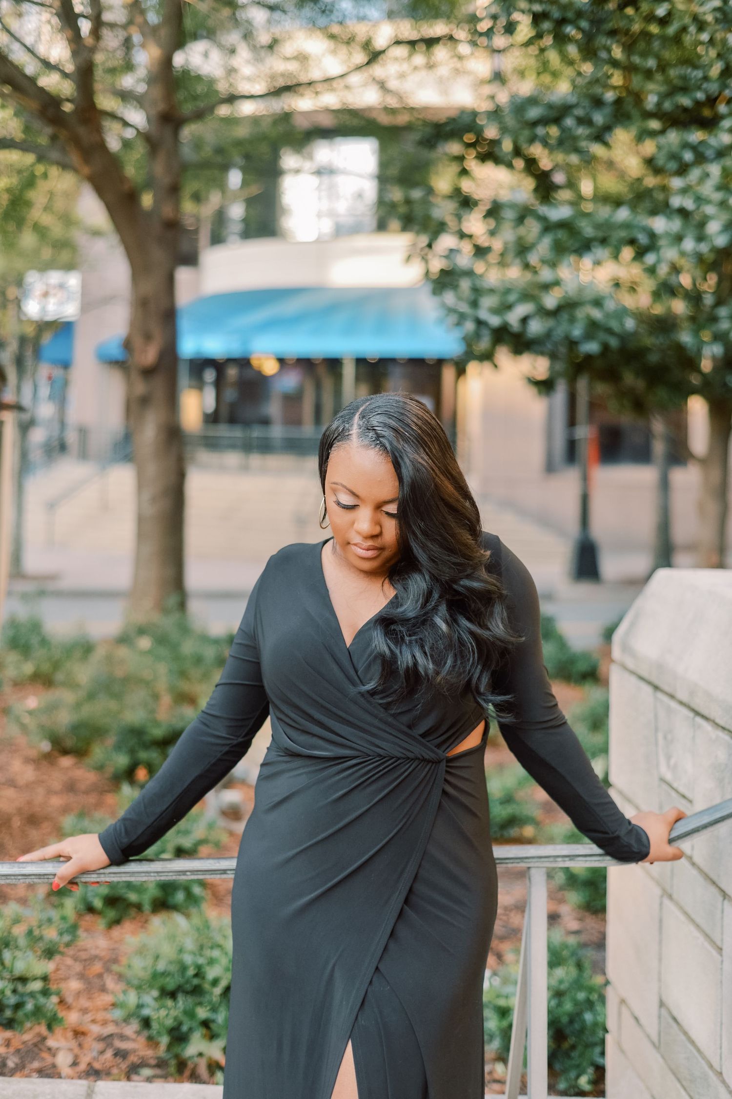 Beautiful woman in elegant black wrap dress with side slit poses on outdoor staircase railing.