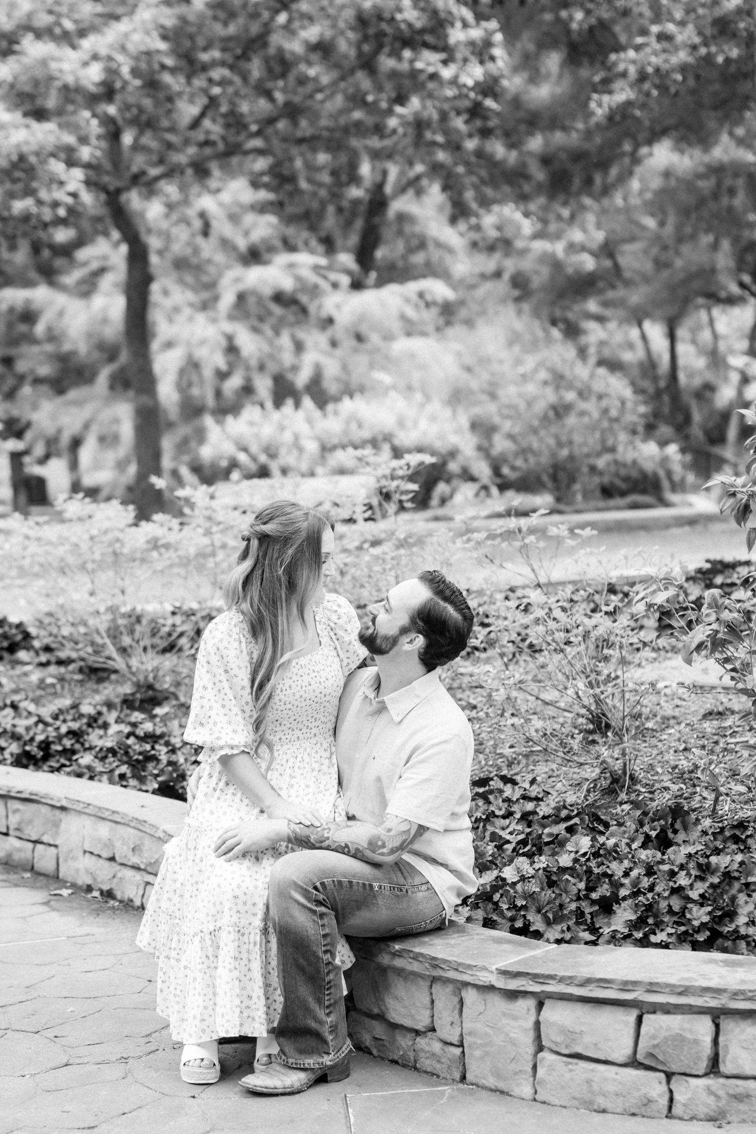Black and white photo of a couple sharing a kiss while seated on a stone garden wall.