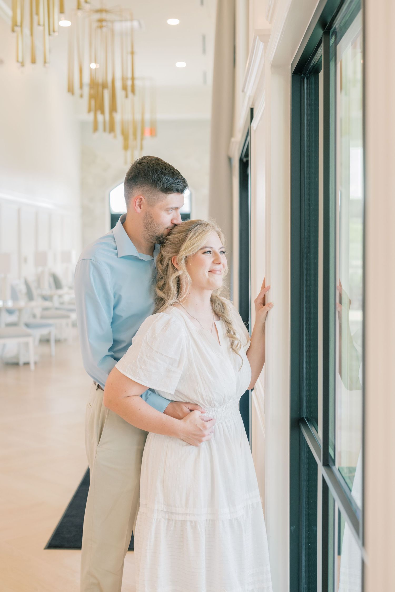Man embracing woman in white dress as they look out large windows in a bright elegant hallway.