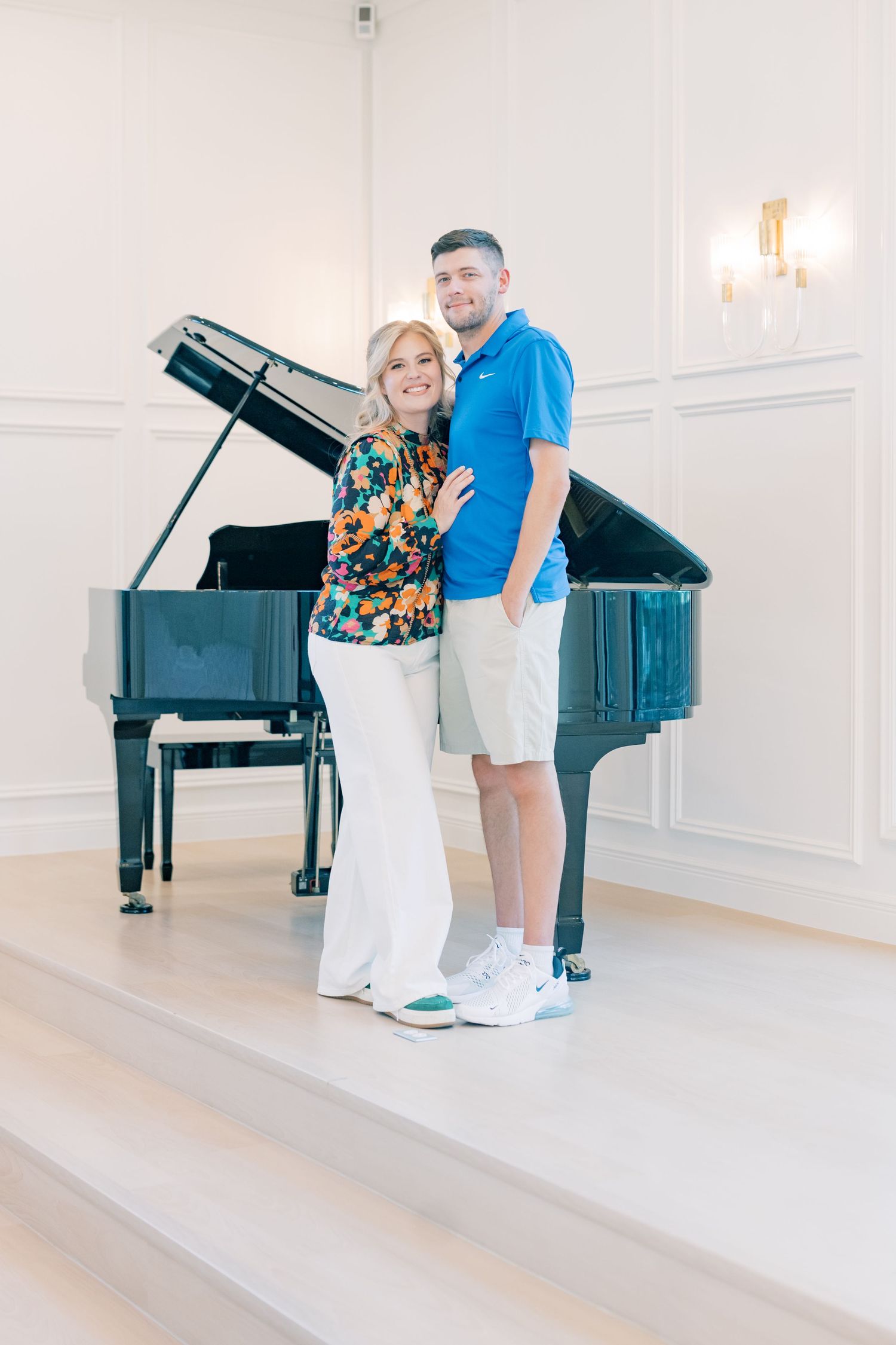 Smiling couple posing together in front of a black grand piano in a bright elegant white room.