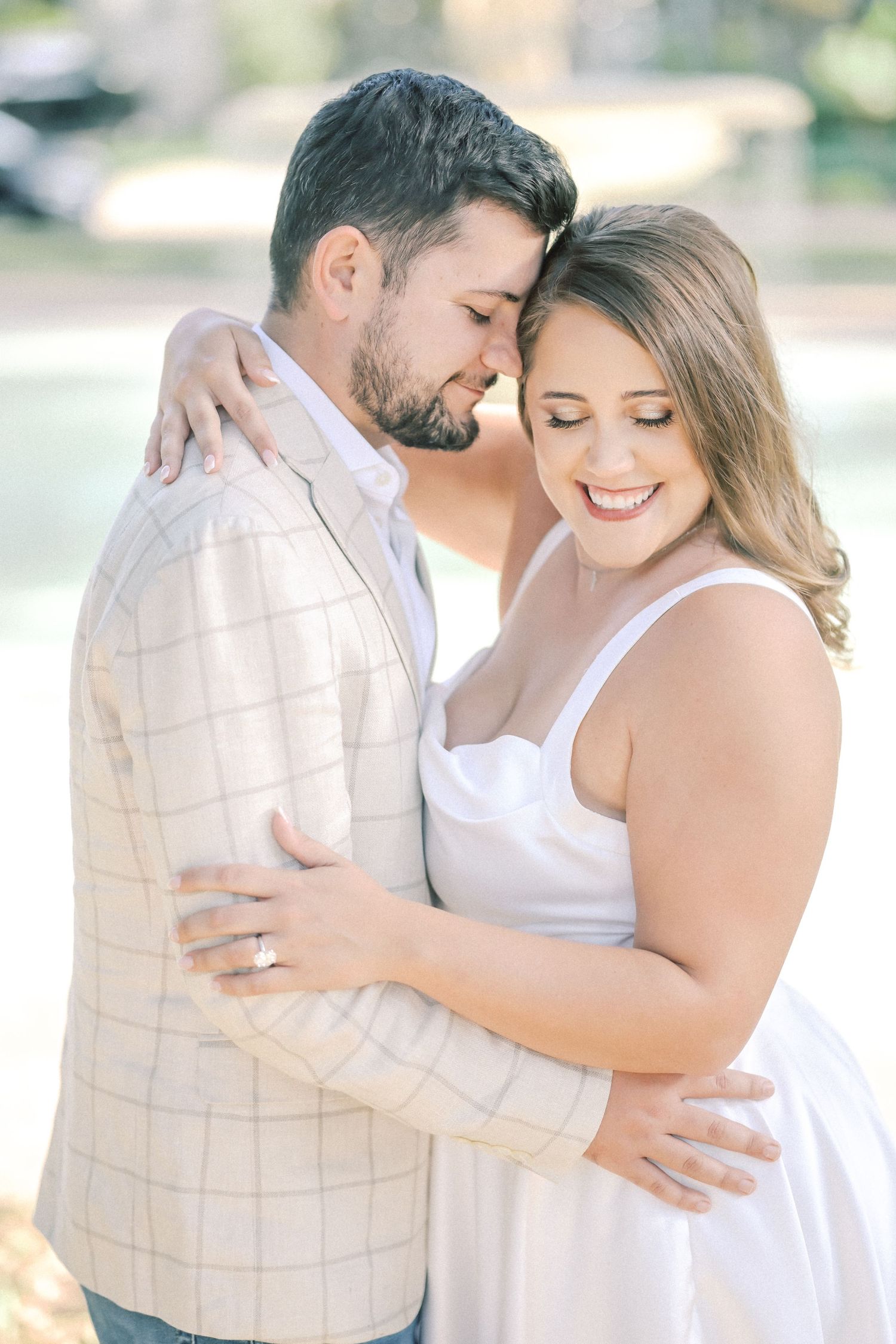 Engaged couple embracing with eyes closed and foreheads touching, the woman smiling and showing her ring in a romantic outdoor portrait.