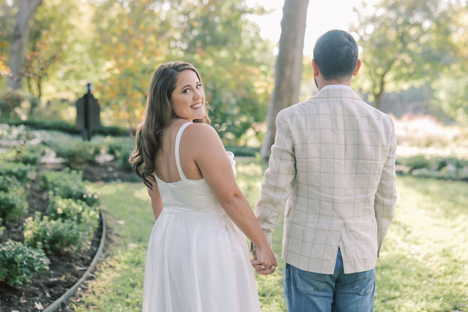 Woman in a white dress smiles back over her shoulder as she holds hands with a man walking through a sunny park.