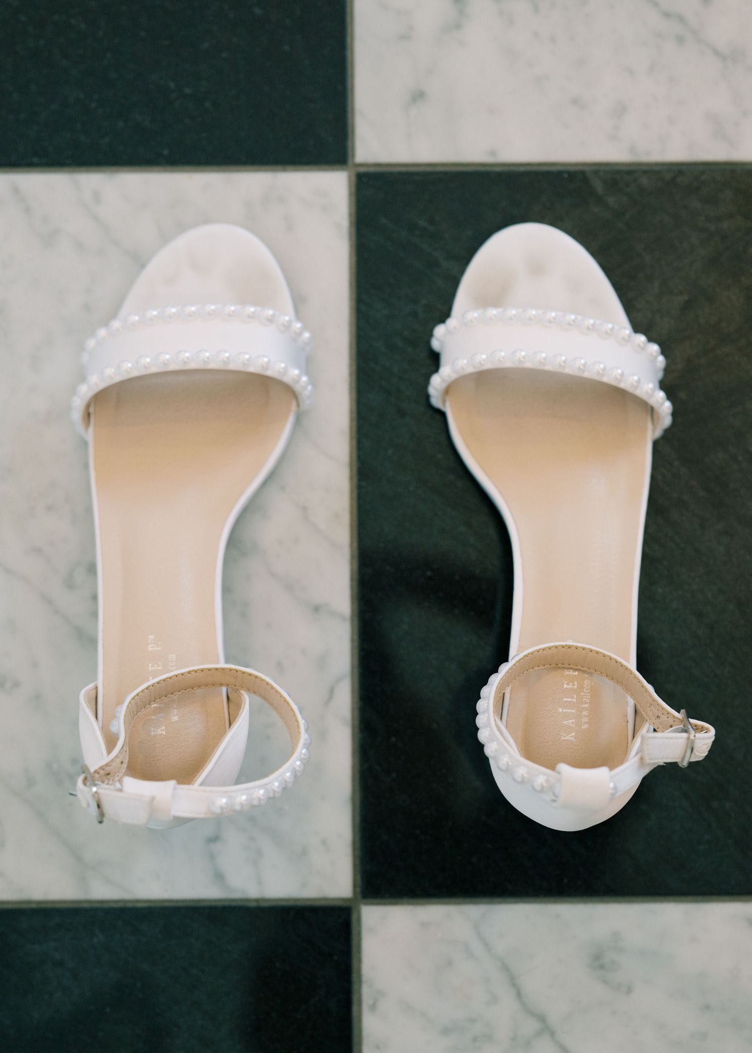 White pearl-embellished bridal sandals with ankle strap displayed on marble and black tile floor.
