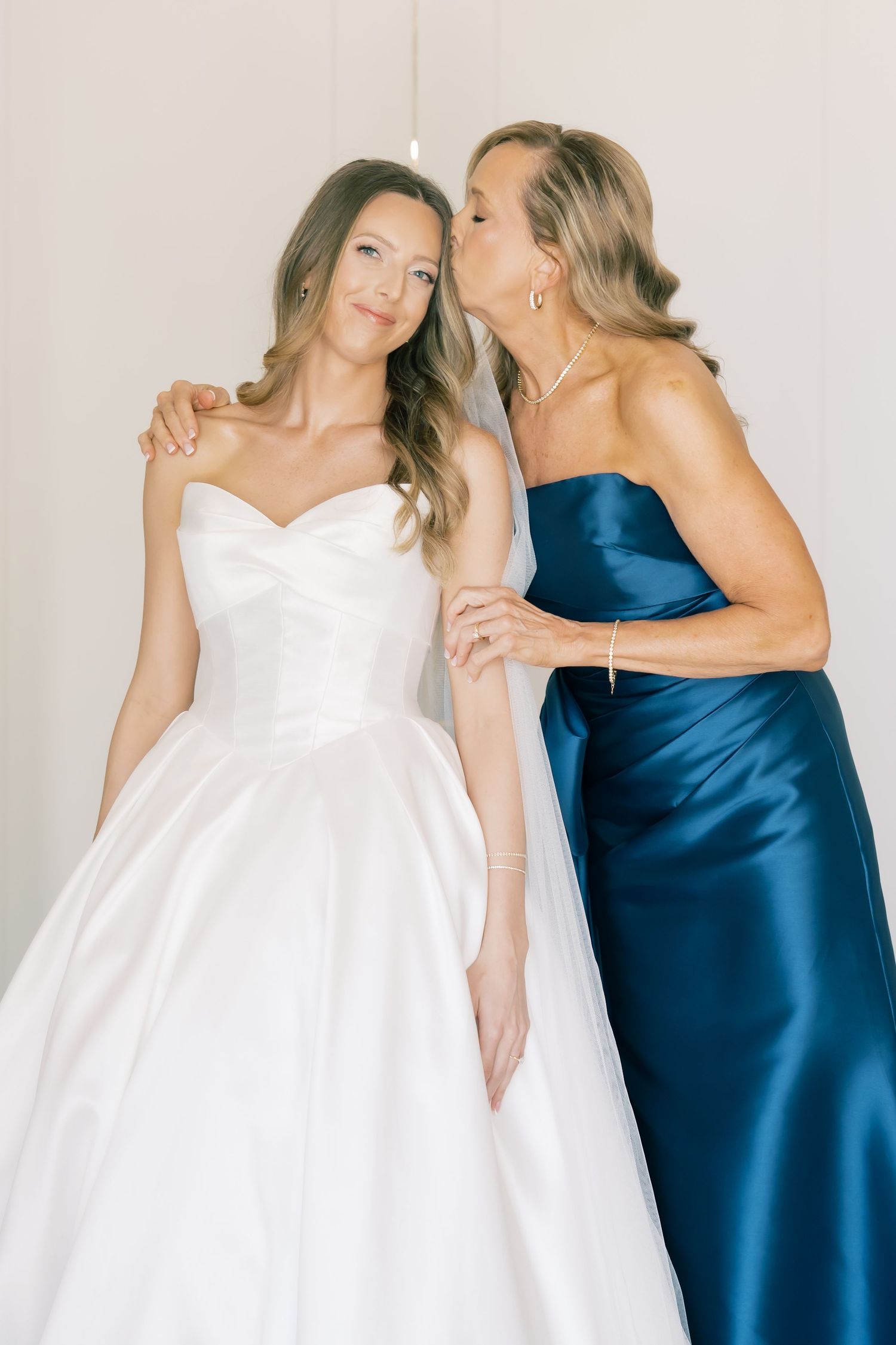 Mother in navy strapless satin gown shares tender kiss on cheek with bride in white ballgown.